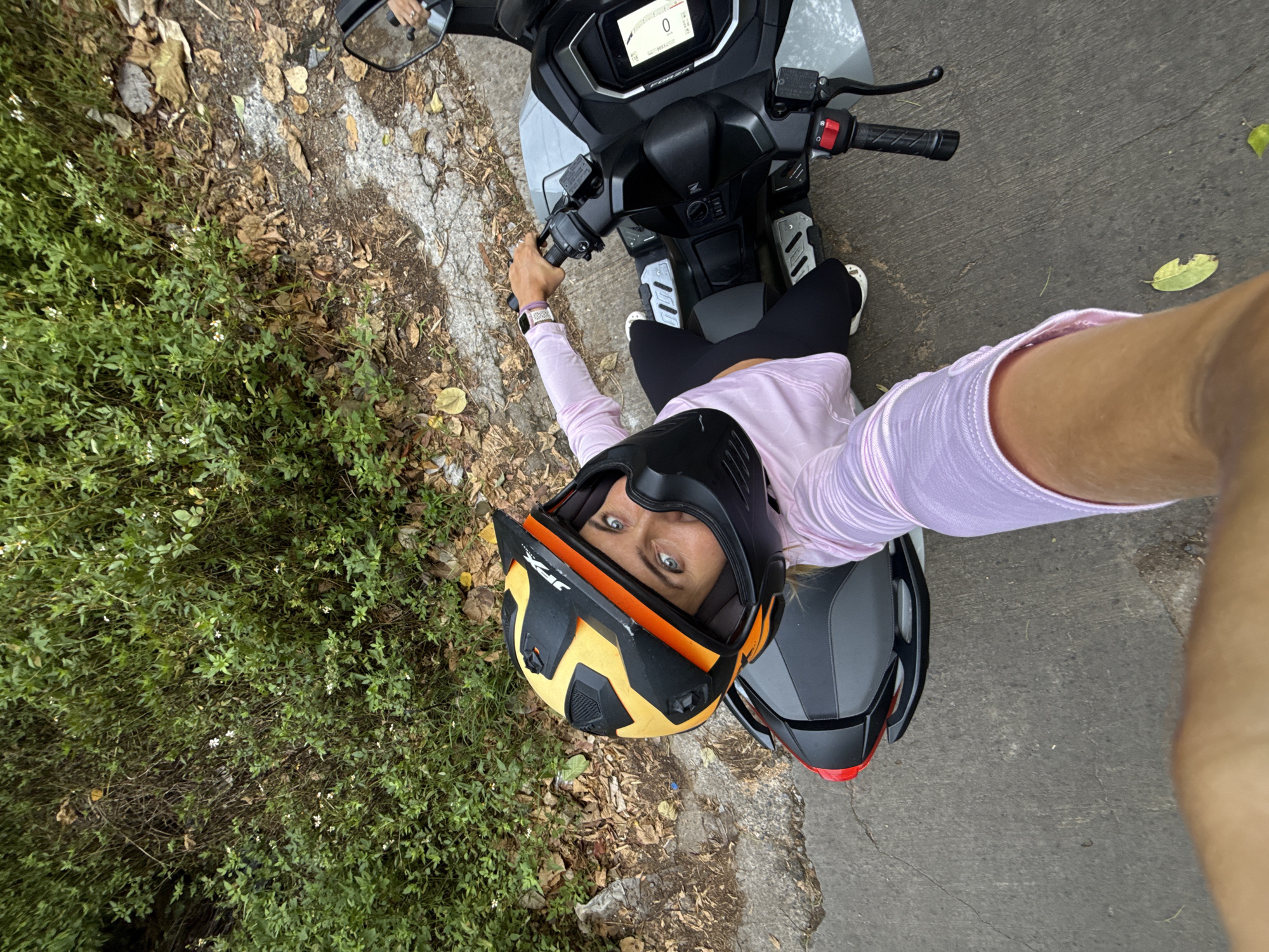 A woman wearing a black and yellow motorcycle helmet and a pink long-sleeved shirt is seated on a grey and black scooter. She is holding the handlebars and looking directly at the camera. The scooter is parked on a paved road next to a patch of overgrown green foliage and fallen leaves. The scene appears to be a roadside in Ko Samui District, Thailand.