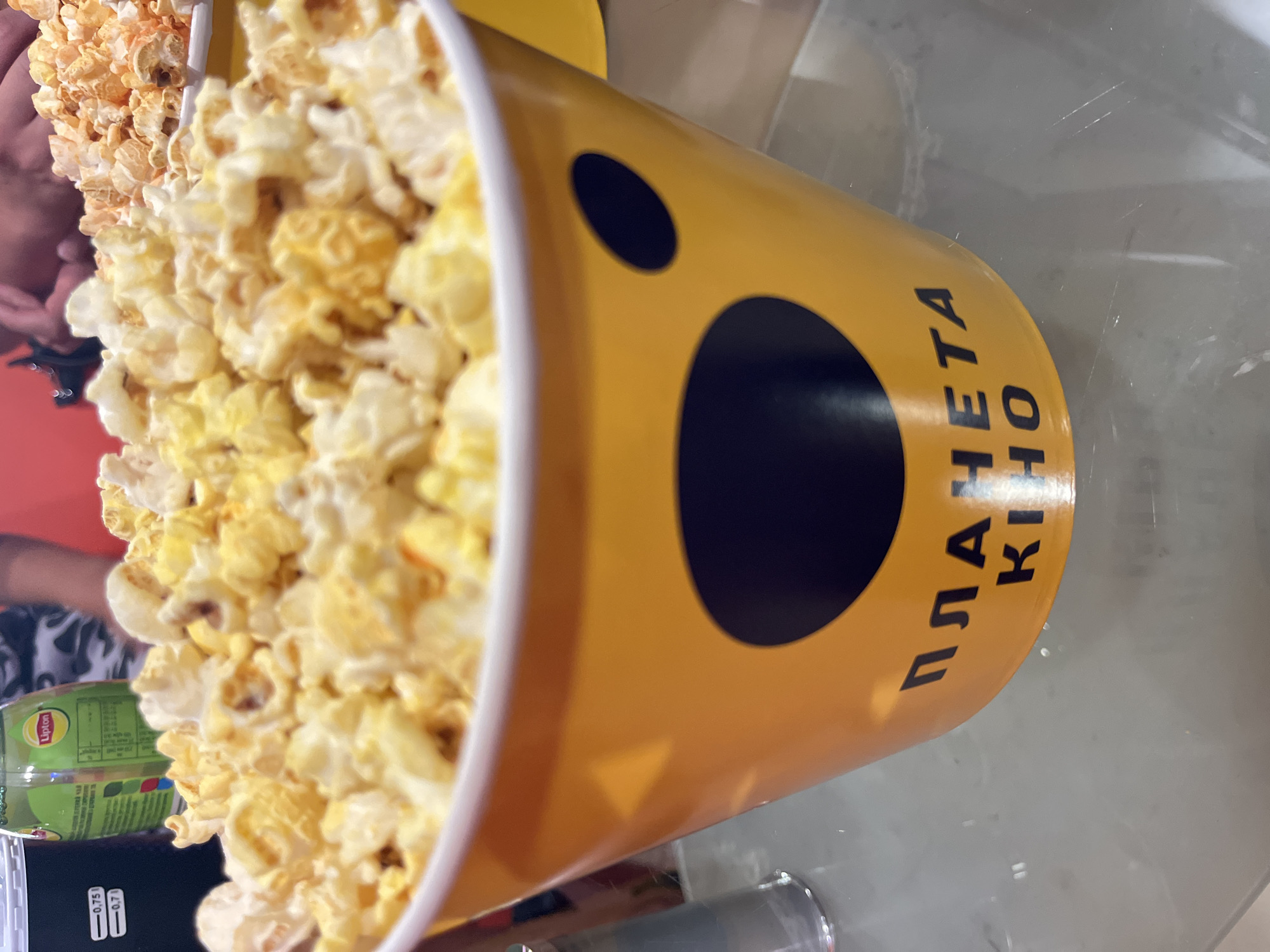 A close-up, eye-level shot shows a large, yellow popcorn bucket filled with popcorn. The bucket has a cartoonish face design with two black oval eyes. Text in black Cyrillic letters is printed on the bucket, reading "ПЛАНЕТА КІНО", which translates to "Planet Kino". This indicates the location is a Planet Kino cinema. A second popcorn bucket, filled with orange-colored popcorn, is visible in the background. The scene appears to be on a glass table.