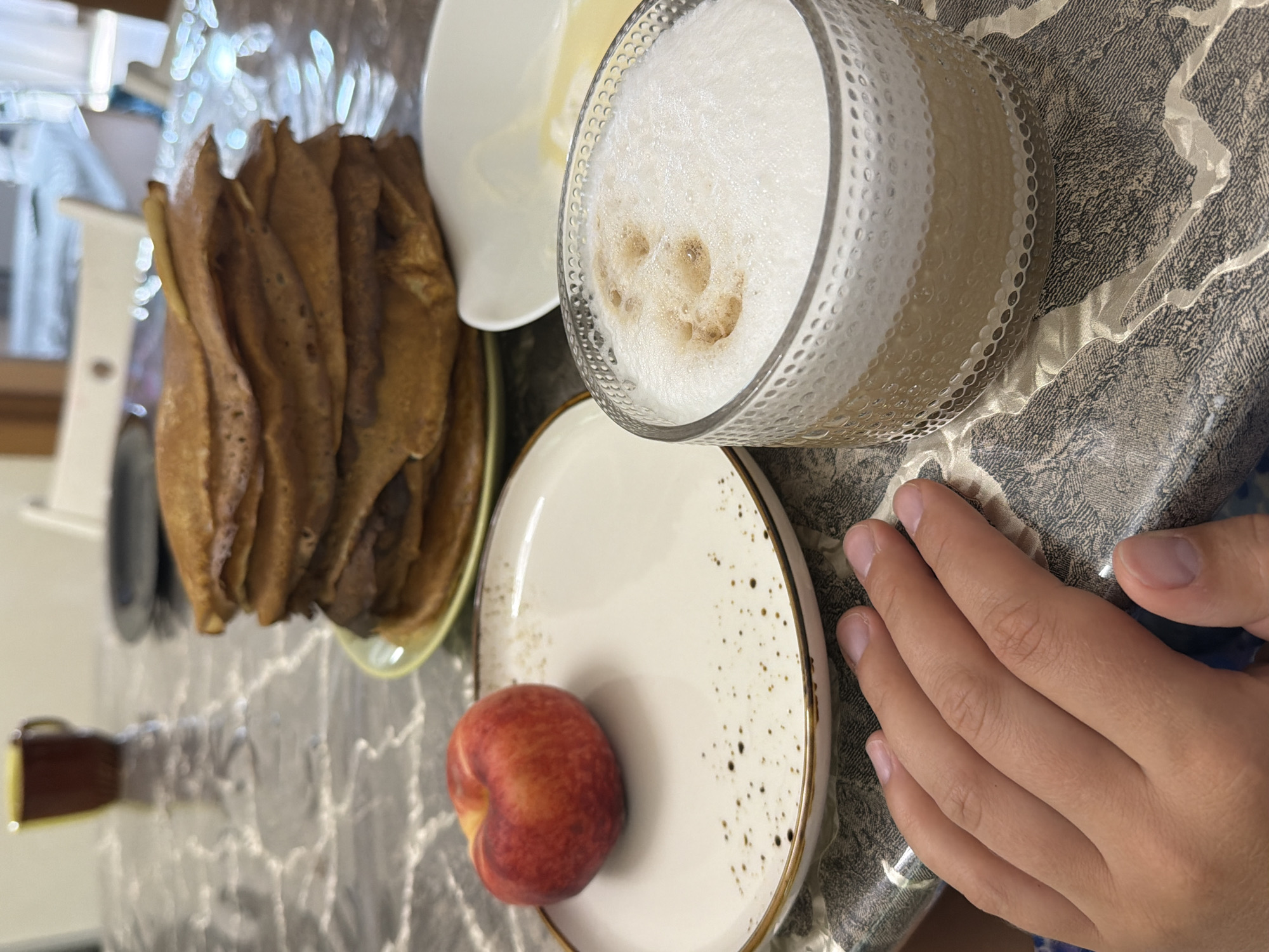 A high-angle medium shot shows a breakfast spread on a patterned tablecloth. In the foreground, a person's left hand rests on the table. To the right of the hand is a clear glass mug filled with coffee and frothed milk. Behind the mug is a white bowl containing what appears to be butter. Further back, a stack of thin pancakes sits on a light green plate. To the left of the pancakes is a white plate with a single peach. In the background, out of focus, is a maroon mug on a reflective surface. The scene is likely indoors, possibly a dining area, with elements suggesting a meal being prepared or consumed. There is no indication of the city of Sochi, Russia, in the image itself.