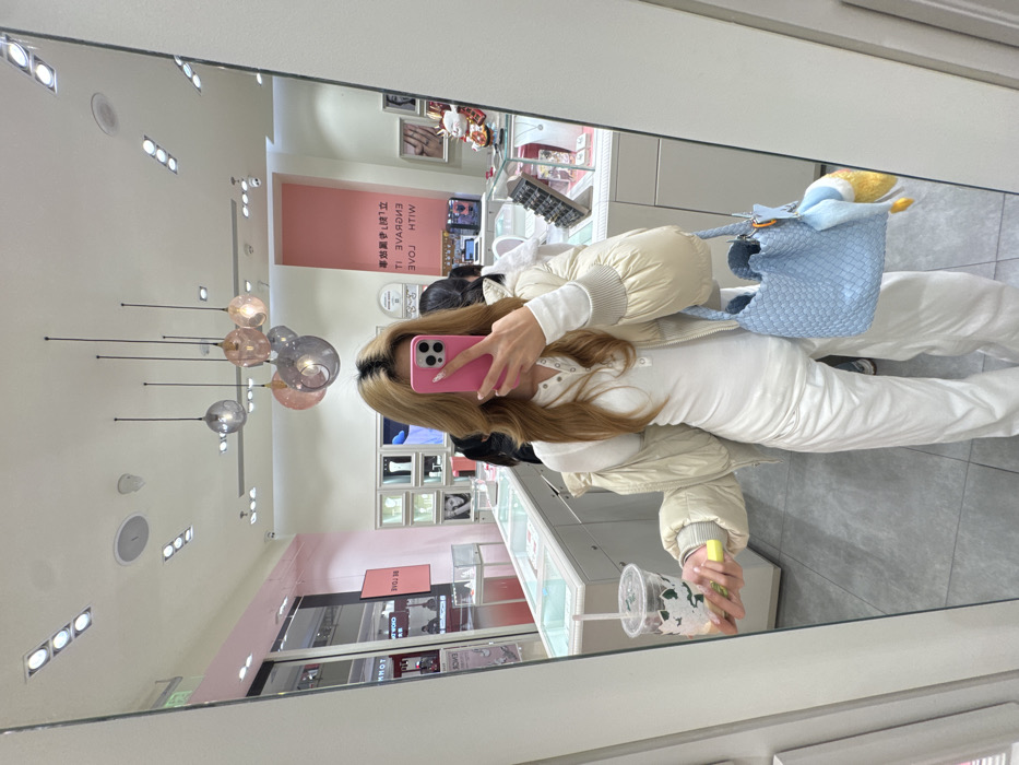 This image captures a mirror selfie taken by a female individual inside a retail establishment, identified as a jewelry or accessory store, likely a Pandora store, situated within a shopping mall in the Yuzhong District of Chongqing, China.
The primary subject is a female with long, light brown hair with visible darker roots, styled with volume. Her face is obscured by a pink smartphone held directly in front of it to capture the image. She is dressed in a white long-sleeved henley-style top with buttons, white loose-fitting pants, and a light beige, puffy bomber-style jacket worn open and draped over her shoulders. Her fingernails exhibit a light-colored polish. In her left hand, she holds a clear plastic cup containing a beverage with ice and a green element, along with a yellow stick-like object. Her right hand carries a light blue woven tote bag, from which a yellow and white cartoon duck plush charm hangs.
The background reveals the store's interior, characterized by white display cases exhibiting various jewelry or accessory items. Pink accent walls are visible, along with multiple modern hanging spherical pendant lights in shades of grey and pink, suspended from a white ceiling that also features recessed spotlights, a security camera, and a white speaker. Visible signage includes "BE LOVE" in black text on a pink background, and partially reflected text reading "ENGRAVE IT WITH LOVE" alongside Chinese characters, further supporting the identification of the store as a Pandora outlet. A white maneki-neko (waving cat) statue with gold accents is displayed on a counter to the far right. In the reflection on the left, a partial "TOMMY HILFIGER" sign indicates an adjacent store within a mall environment. Other individuals are partially visible as blurred reflections in the background, suggesting the presence of other customers or staff.