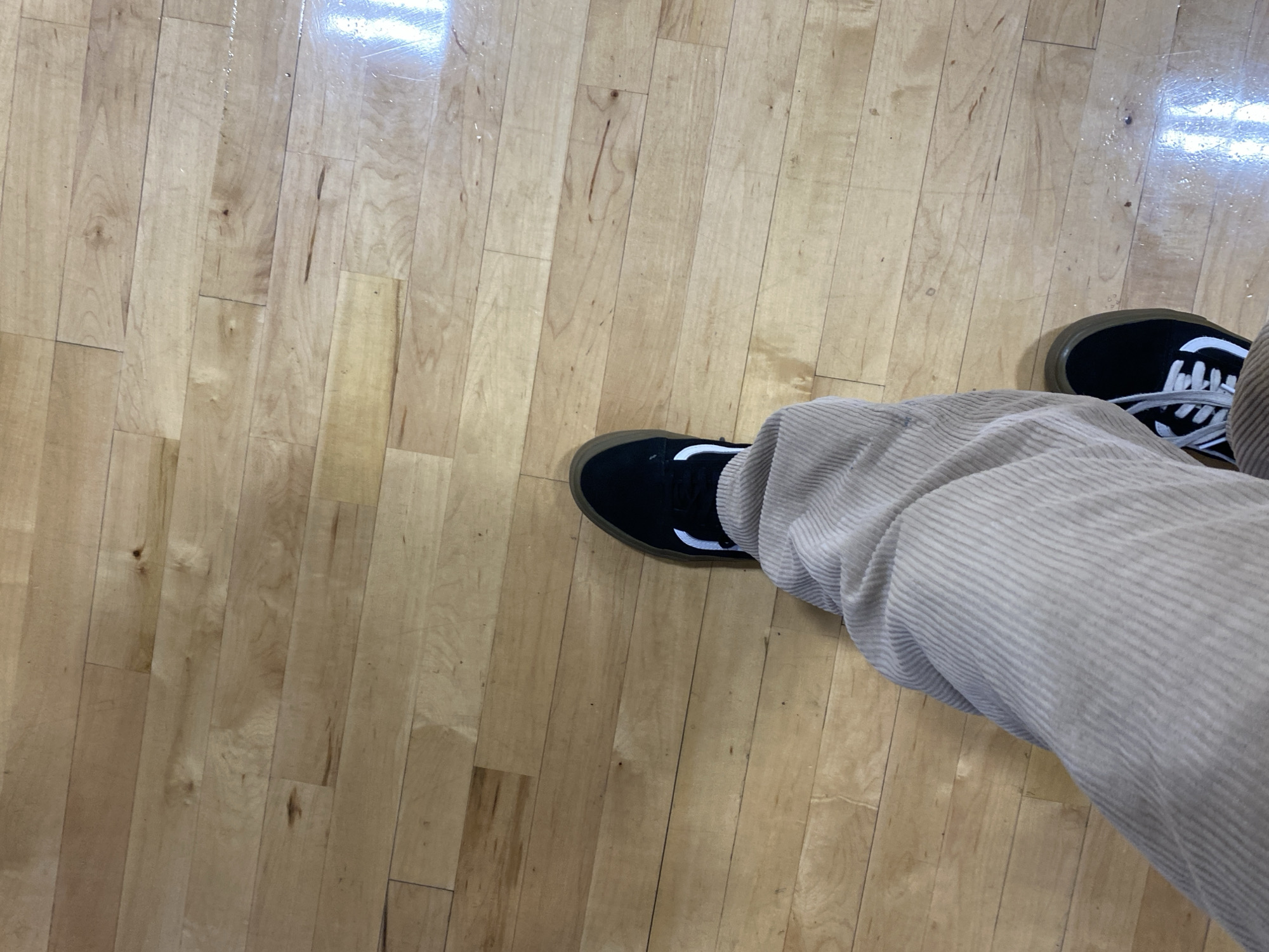 The image shows a person's legs and feet standing on a light-colored wooden floor. The person is wearing light brown corduroy pants and black sneakers with white laces and a white stripe. The floor appears to be made of planks and has a glossy finish, with some reflections visible. The scene is indoors and is devoid of any distinguishing features that would indicate a specific location in Virginia Beach, United States of America. There are no people other than the legs and feet visible, and no significant interactions are depicted. The environment is a plain wooden floor.