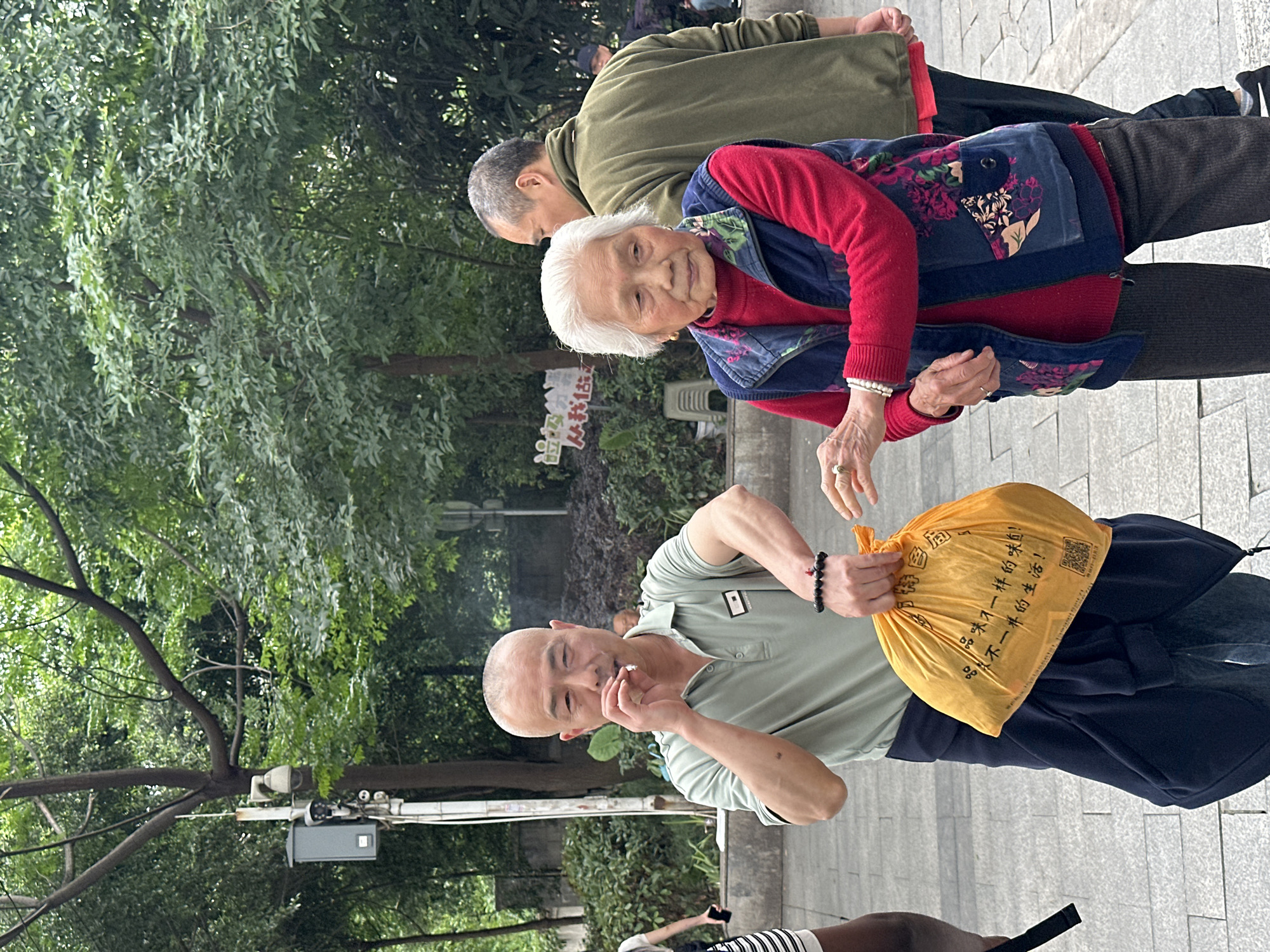 The image shows three people and some greenery in what appears to be a public outdoor setting. In the foreground, a bald man in a light green polo shirt holds a cigarette near his mouth with his right hand and a yellow plastic bag with Chinese writing on it in his left hand. The text on the bag includes the phrases "品味不一样的味道!" and "品不一样的生活!".
Behind him, an elderly woman wearing a red sweater and a blue floral vest looks towards the camera. Her right hand is extended as if reaching for something. Further behind her is an older man in an olive-green jacket. The environment includes trees, bushes, and a gray tiled surface, which suggests a sidewalk or plaza. The presence of security cameras mounted on a pole to the left suggests it is in the city of Shapingba District, China.