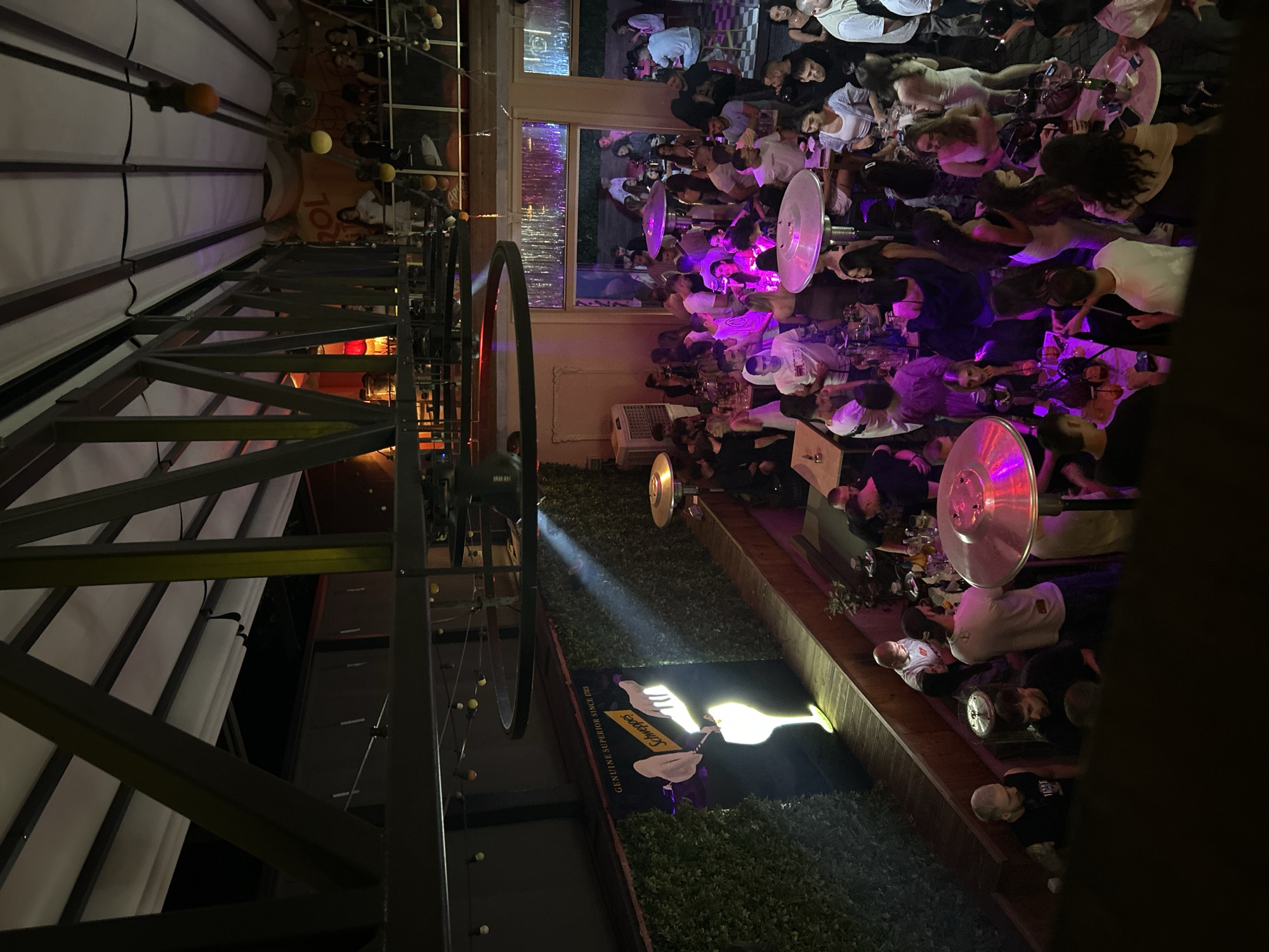 The image depicts a crowded outdoor seating area of a bar or restaurant, likely at night, in Plovdiv, Bulgaria. The camera angle is high, possibly from an elevated position. The area is lit with a combination of ambient lighting and purple-tinted uplighting, creating a lively atmosphere. Numerous people are seated at tables, socializing and drinking. Patio heaters provide warmth and light. A decorative metal structure with bicycle wheels is a prominent feature. A Schweppes advertisement is visible near a planter.