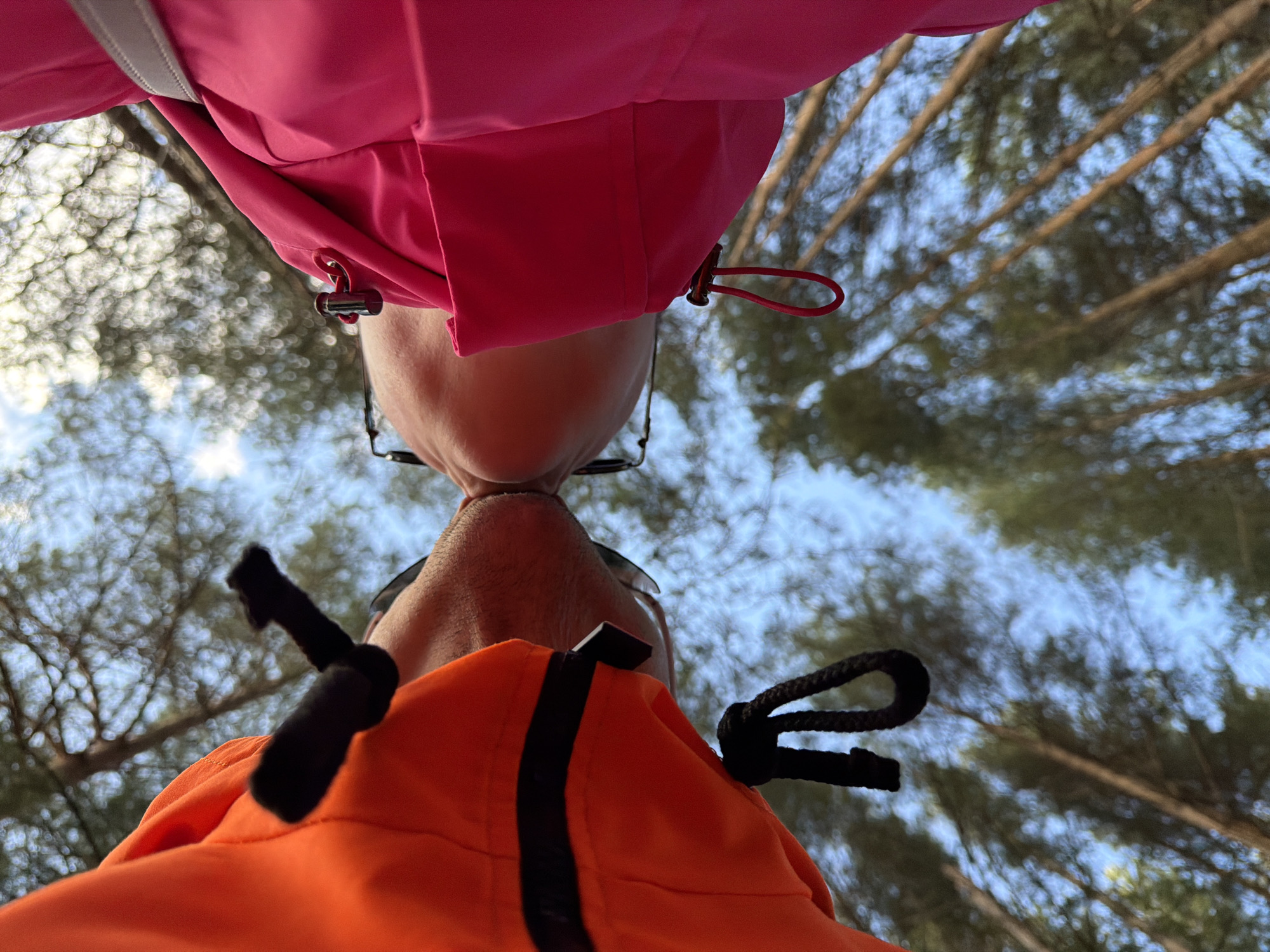 A low-angle shot depicts two individuals standing close together, noses touching, against a backdrop of trees and sky. The person on top wears a pink jacket, and the person below wears an orange jacket. Both are wearing glasses. The photo appears to be taken outdoors, possibly in a wooded area. While the exact location in Chaikovsky, Russia, cannot be definitively determined from the image alone, the presence of coniferous trees suggests a forest or park setting common to the region.