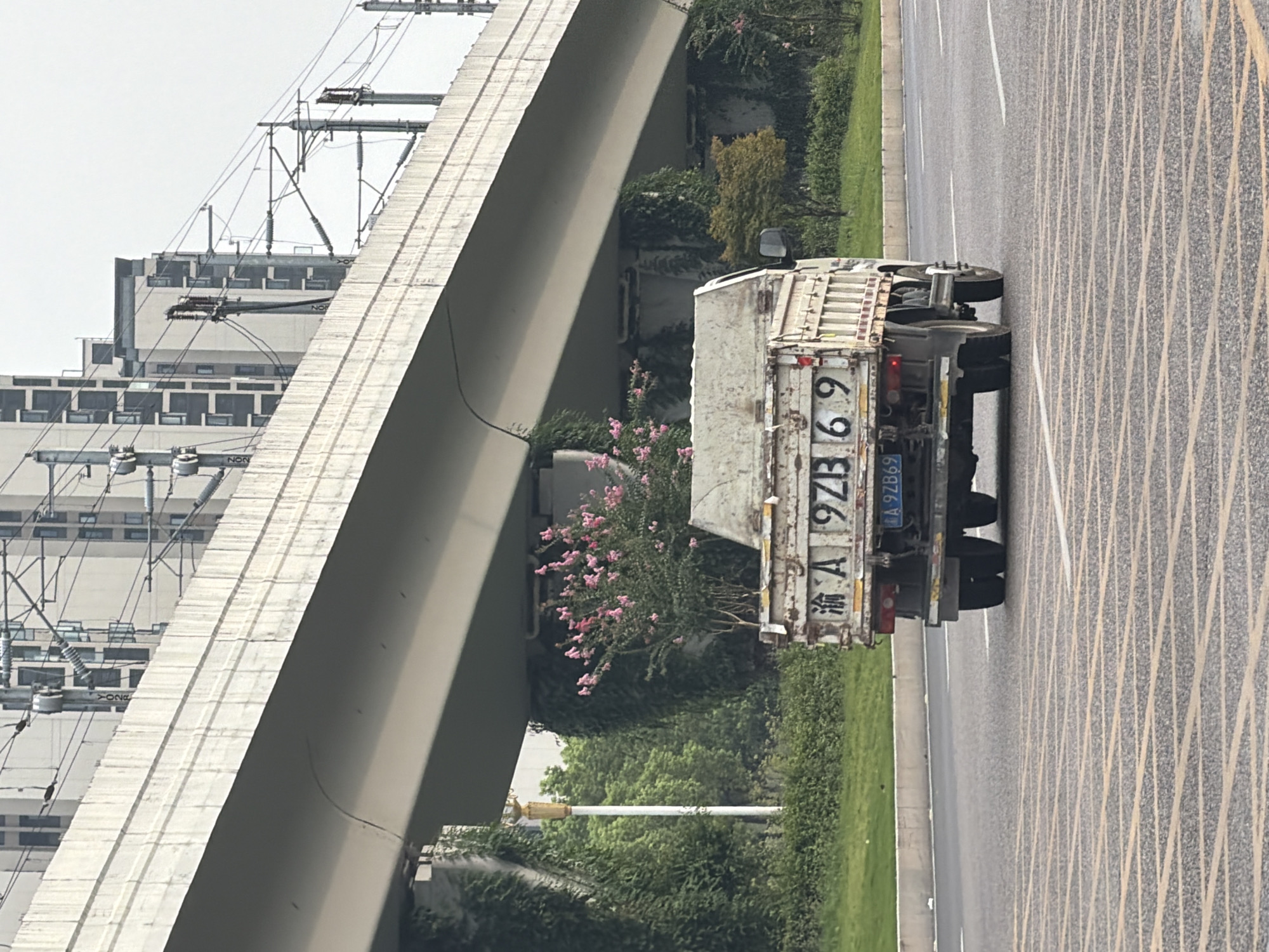 A white dump truck with a license plate that reads "渝A 9ZB69" is driving on a road. The truck is seen from behind and slightly to the side. The truck bed is empty and appears to be made of metal. In the background, there is a large concrete overpass with train tracks on top. Buildings and trees are visible behind the overpass. The road has yellow and white markings. The scene appears to be in an urban or suburban area.