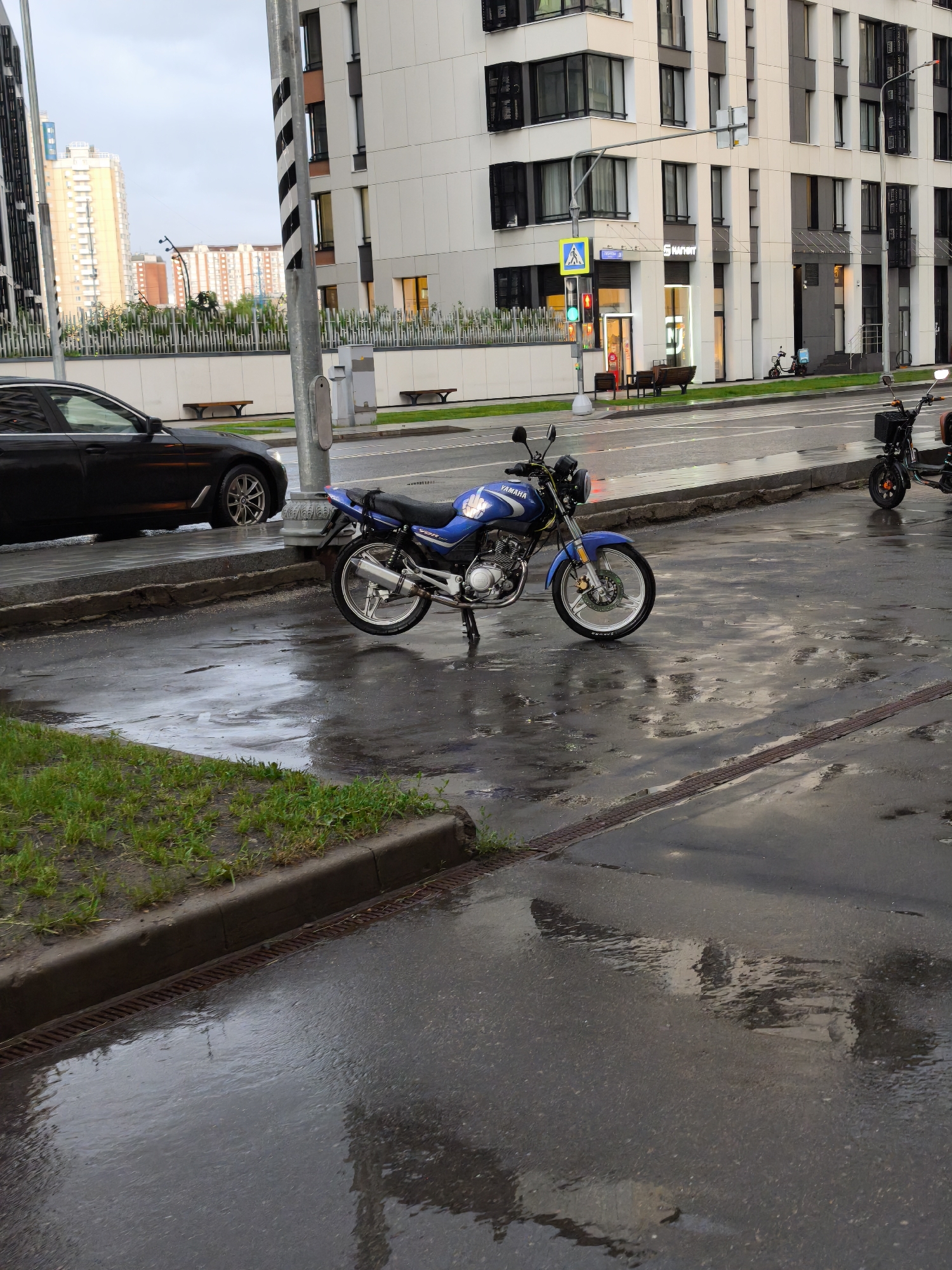The image depicts a street scene in Moscow, Russia on a wet day. A blue Yamaha motorcycle is parked on the side of the road near a curb and grassy area. A black car is parked further down the road next to a light pole. In the background, modern apartment buildings are visible. A pedestrian crossing sign and a traffic light can be seen. In the distance, other high-rise buildings are visible, suggesting a densely populated urban area. Another small motorbike is parked further ahead. The wet road surface reflects the surrounding environment.