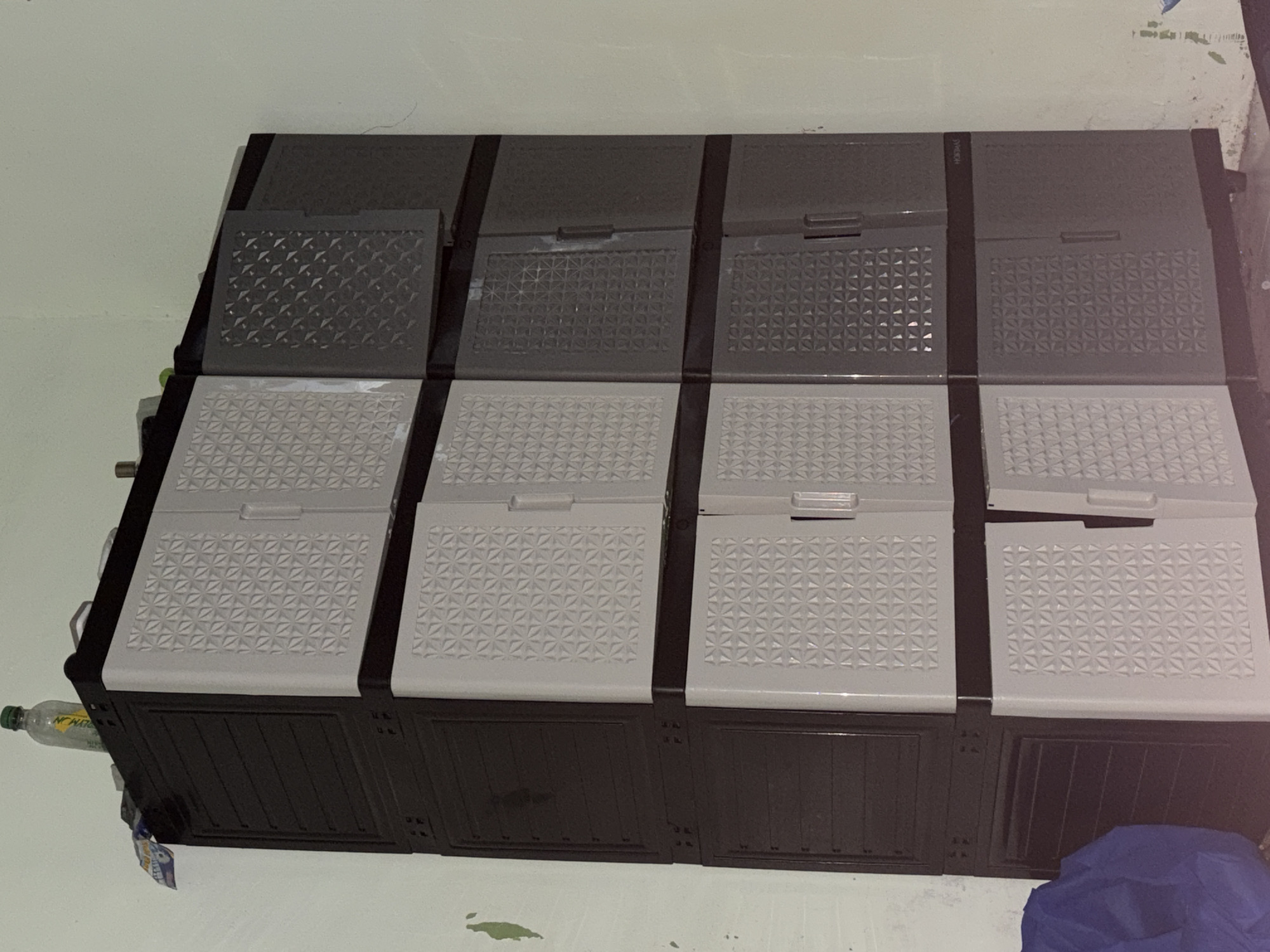 A tall, multi-tiered plastic cabinet stands in a corner against a light green wall in Tupah, Indonesia. The cabinet is dark brown on the left and alternates between light gray and dark gray on the right, with each tier having two doors. The light gray doors have a raised geometric pattern, while the dark gray doors have a darker, embossed geometric pattern. The top of the cabinet holds a clear plastic bottle with a green cap and a yellow label, along with other unidentifiable items. Below the cabinet, a folded blue cloth is visible on the floor. The wall shows signs of wear, with patches of peeling paint.