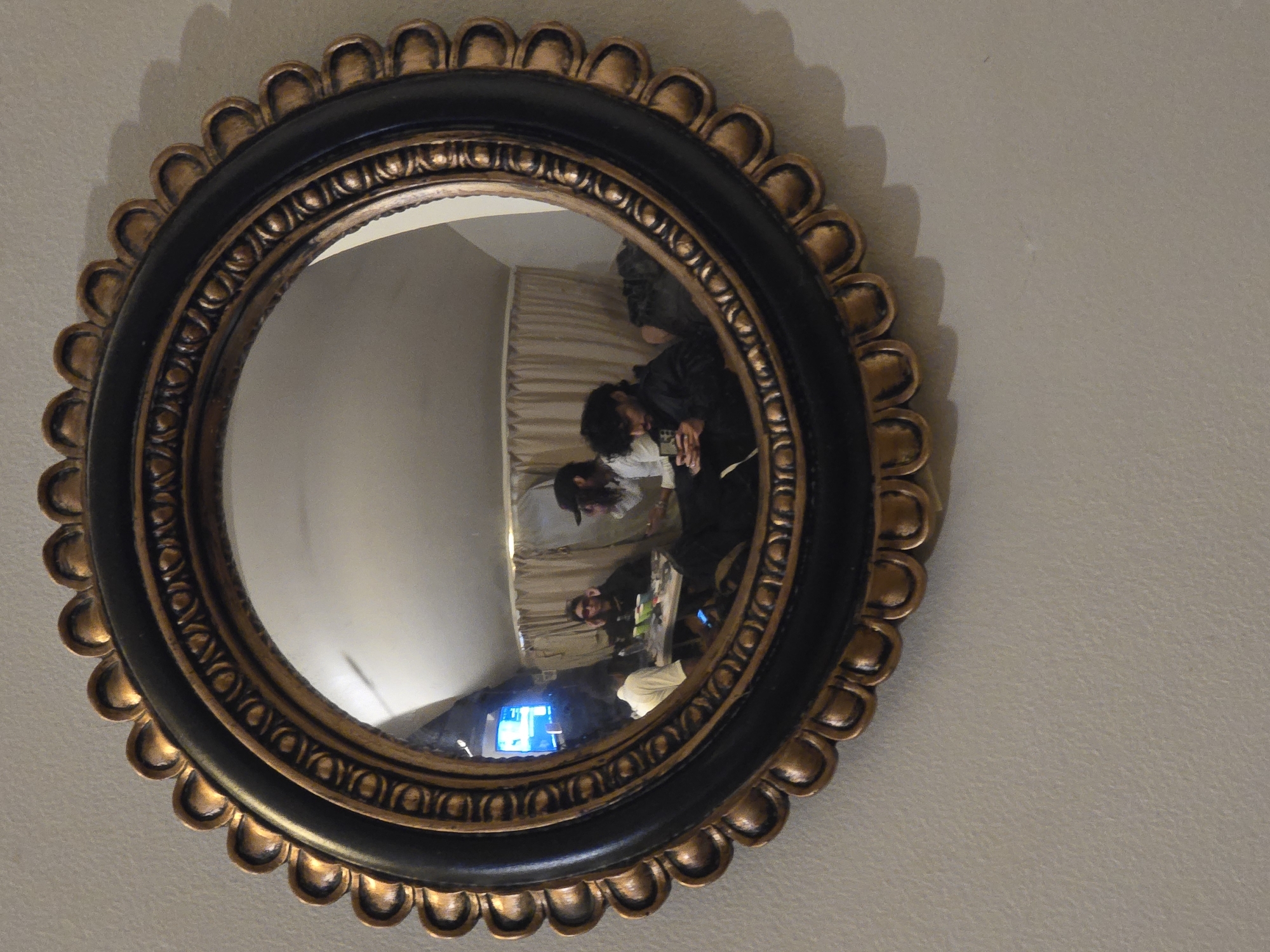The image shows a round mirror with a black and gold frame hanging on a light beige wall. The frame has a decorative design of repeating scallop-like shapes around its outer edge, followed by a pattern of raised ovals around the inner edge. Reflected in the mirror is a room with a light-colored curtain. Several people are visible in the reflection: one person in a black jacket is bending over a table; another person in a white shirt is looking at something on the table; two other people can be seen in the background. A blue-lit screen is also visible in the reflection. The scene is not identified as being in Paris, France.