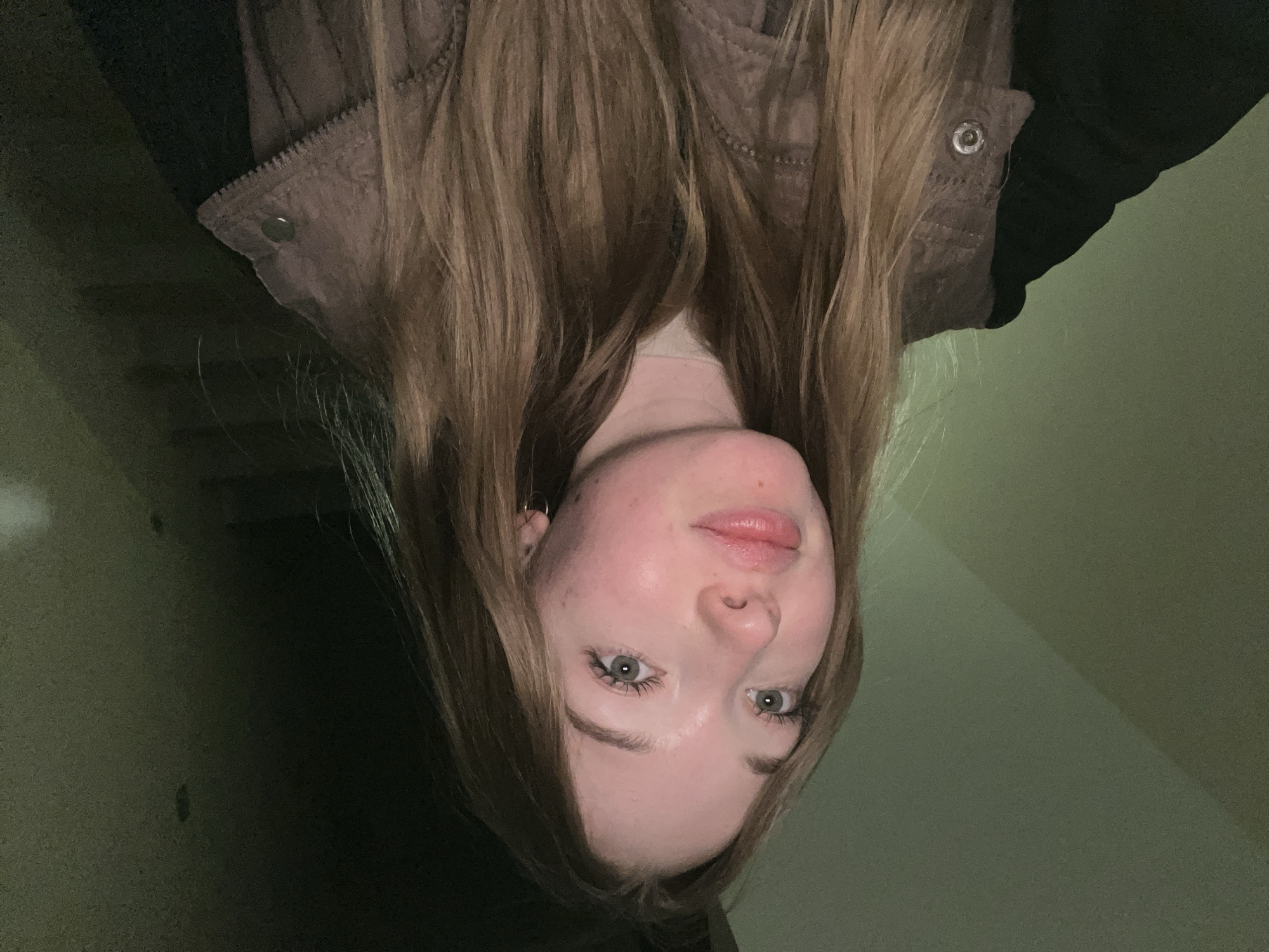 An upside-down medium shot shows a person with long, straight, light brown hair. The person is wearing a brown jacket over a black top. The background appears to be a wall in an olive green color, possibly in the city of Chaikovsky, Russia. The person’s features are visible, including light eyes, a small nose, and light pink lips.