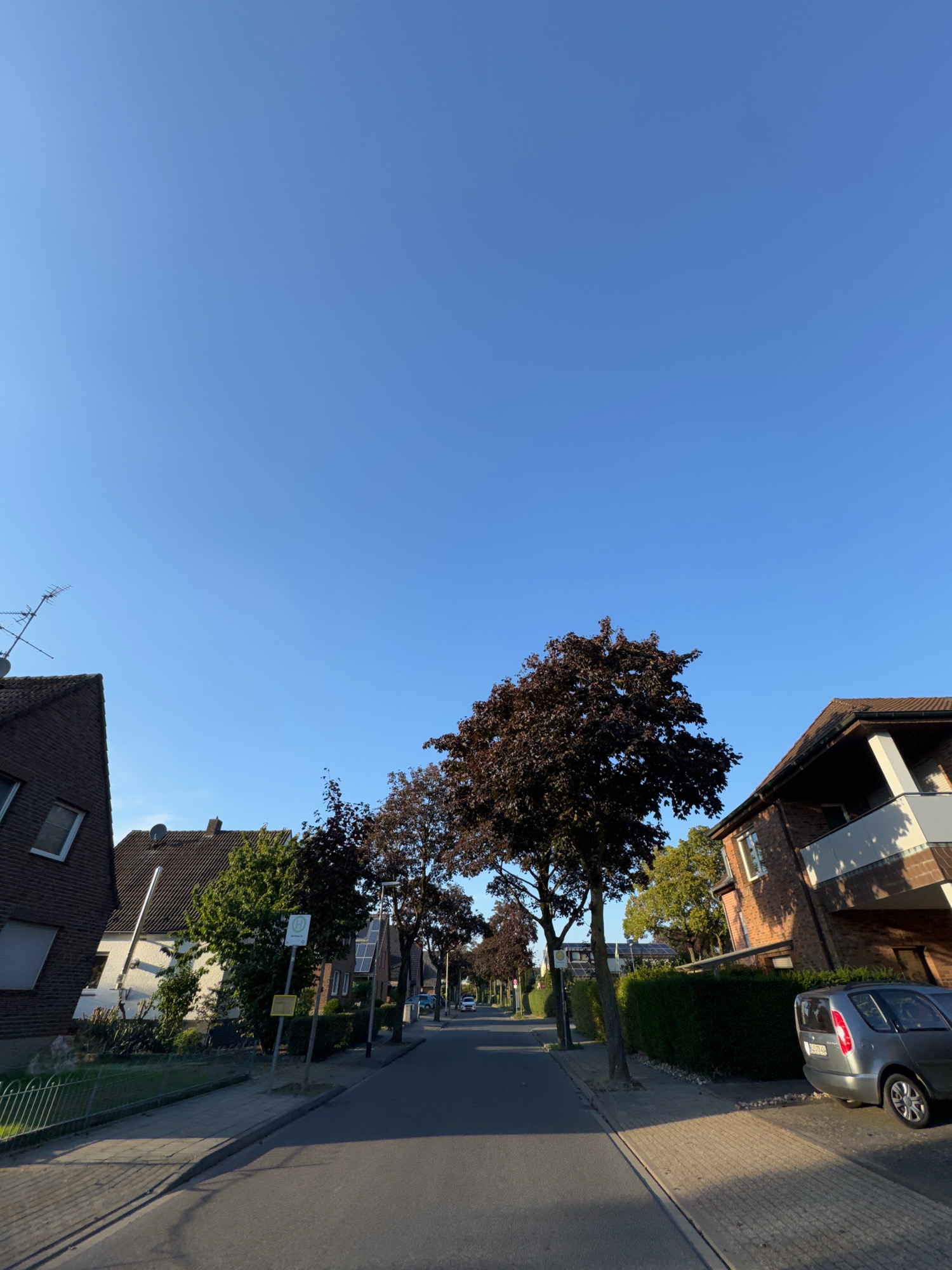 The image depicts a street scene in Cleves, Germany, likely a residential area. The sky is clear and blue. On the left side of the street, there are two houses made of dark brick. The house closest to the viewer has an antenna on its roof. Further down the street on the left, there is a white house with solar panels on its roof. There are trees lining the street, some with dark red leaves. On the right side of the street, there is a multi-story building with a balcony and a car parked in front of it. A silver station wagon is parked on the right side of the street. A bus stop sign is visible on the left side of the street. The street is paved with asphalt, and there are sidewalks made of brick pavers. The scene appears to be during daylight hours with sunlight casting shadows.