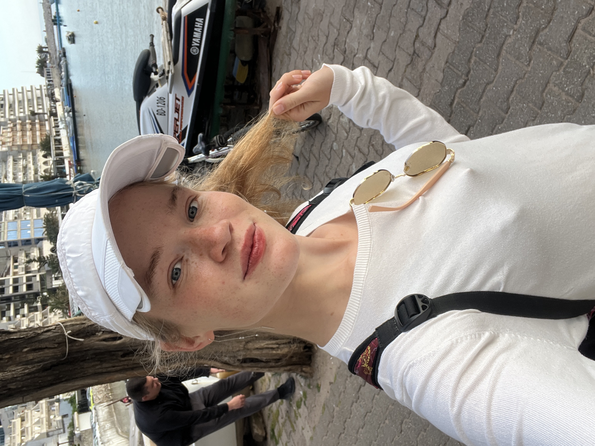A young woman is the focus of this slightly angled, waist-up selfie. She is wearing a white long-sleeved shirt and a white baseball cap, with round sunglasses resting on her chest. She has blonde hair and freckles, and she is holding a strand of her hair.
In the background, a Yamaha personal watercraft is visible, as well as a marina and some buildings. Below, a man is sitting on a bench, near the root of a tree. The pavement of the surrounding area is made up of interlocking stones.
The setting is Boreti, Montenegro, as evidenced by the appearance of the marina, architecture, and vegetation. The woman appears to be enjoying a day outdoors.