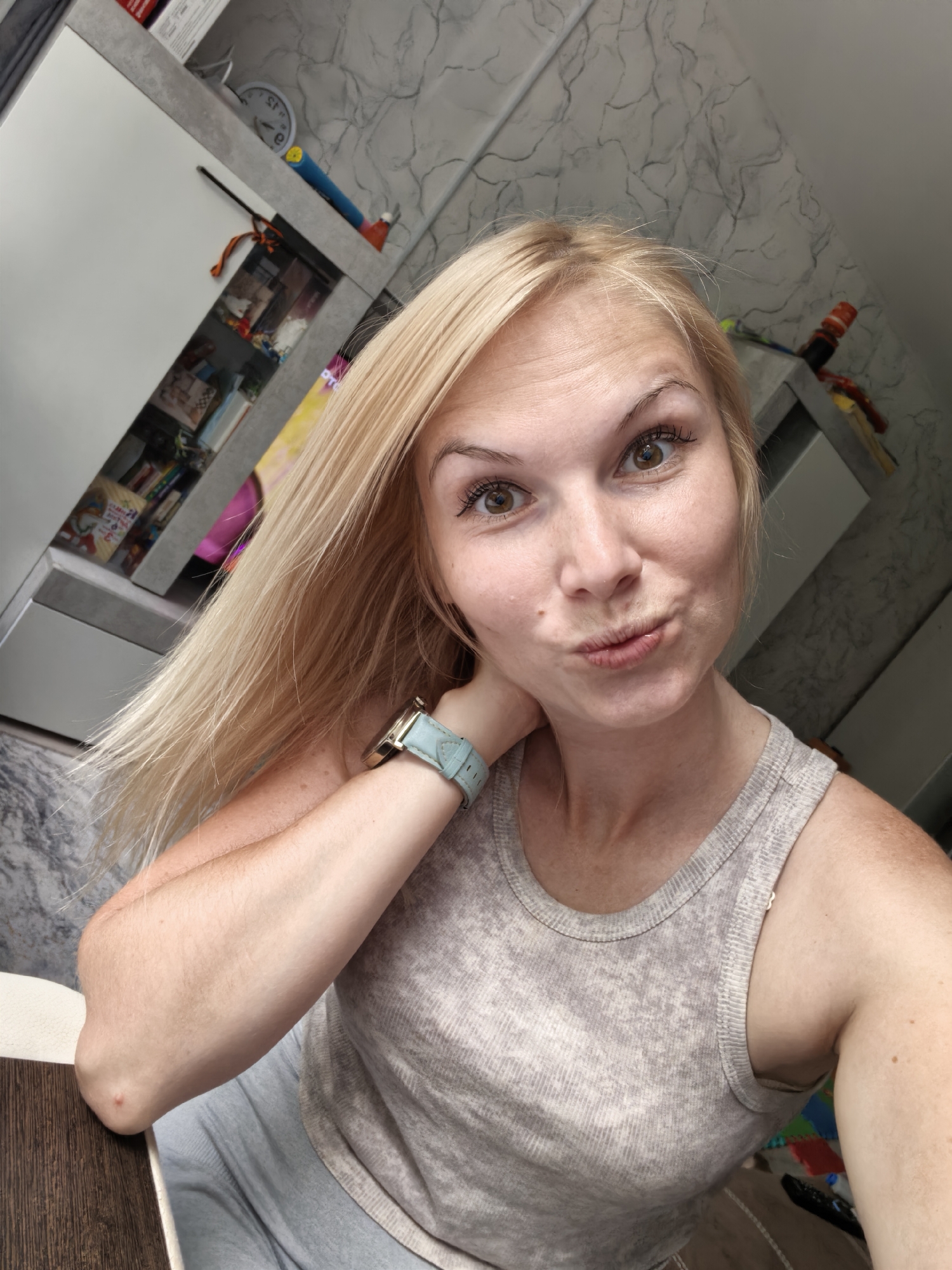 A selfie is taken from a high angle of a Caucasian woman with blonde hair and brown eyes. She is wearing a light gray tank top and a light blue watch on her left wrist. Her right hand is behind her neck, and her lips are puckered. The background appears to be an indoor setting with shelves filled with various items, including books and toys. The wall has a marbled gray and white pattern. The flooring is a dark wood laminate. There are some colorful play mats visible on the floor. The overall lighting suggests it is daytime. The image does not provide any specific information about the location being Sterlitamak, Russia.