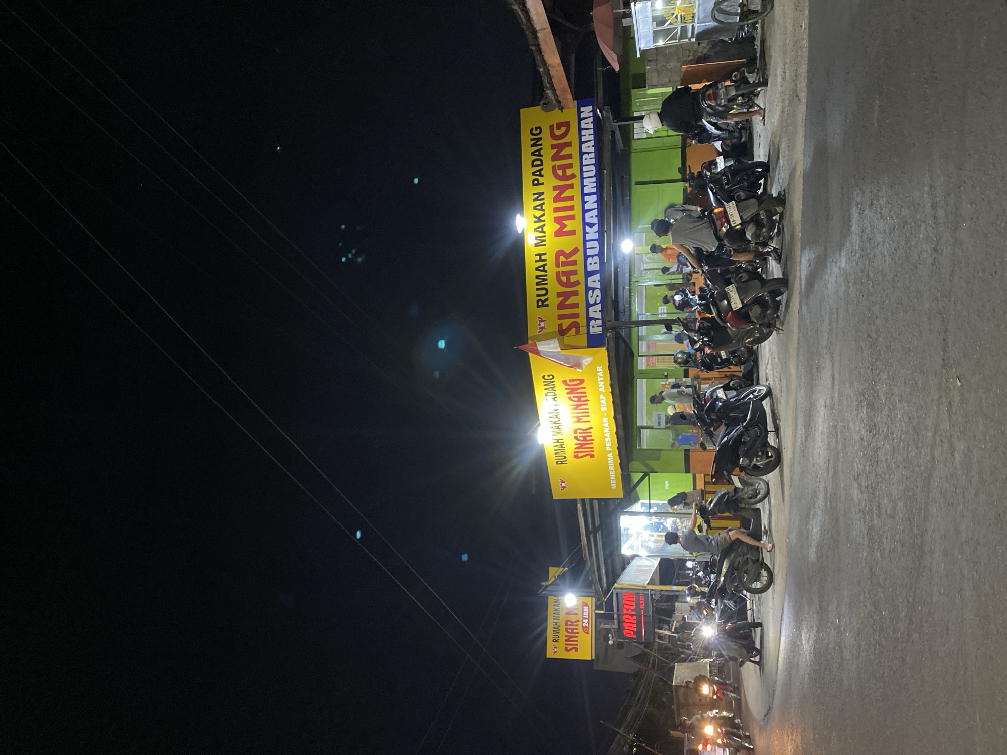 This image captures a street scene at night in the city of Kasihan, Indonesia, specifically in front of "Rumah Makan Padang Sinar Minang" which translates to "Sinar Minang Padang Restaurant". The restaurant features prominent yellow signs with red and blue lettering. The sign on the left reads "SINAR M" and below it "24 JAM". The larger sign on the right states "RUMAH MAKAN PADANG SINAR MINANG RASA BUKAN MURAHAN", meaning "Sinar Minang Padang Restaurant, Taste Not Cheap".
The street is busy with parked motorcycles, indicating a popular establishment. Several people are visible, some standing and others seated on or near their motorcycles. A few individuals are inside the brightly lit restaurant. The environment is urban, with power lines visible overhead against the dark night sky. The overall scene depicts a lively evening at a local eatery, a common sight in Indonesian cities.