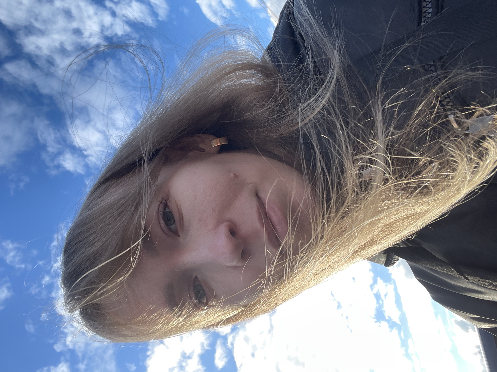 The image is a selfie taken from a low angle, looking upwards at a woman's face and the sky. She has light brown hair and fair skin. She is wearing a dark-colored jacket. The background shows a blue sky with scattered white clouds. The photograph was likely taken outdoors during daylight. The city location (Perm, Russia) cannot be confirmed based on the visual information in the image.