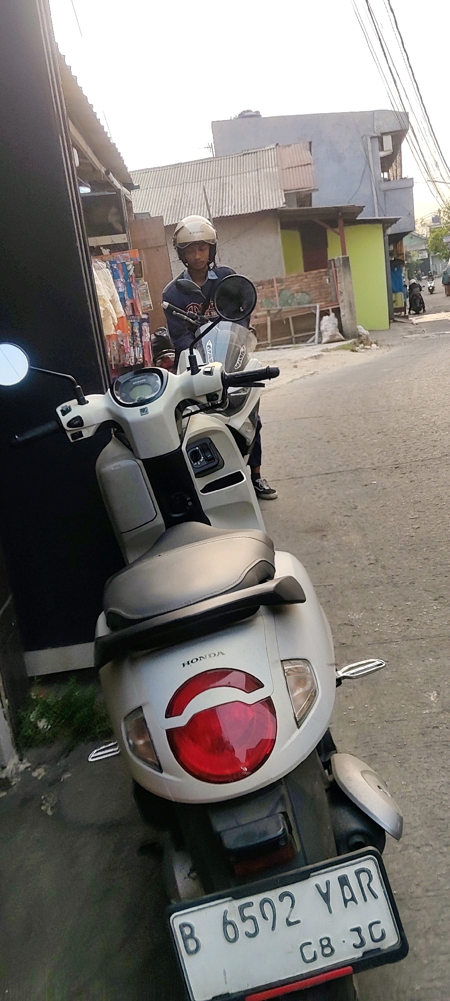 In Mekarmukti, Indonesia, a person wearing a light-colored helmet and a dark-colored jacket stands next to a light-colored scooter on a paved street. The scooter is parked on the left side of the image, with its front facing away from the viewer. The scooter's license plate reads "B 6592 YAR C8 3C". The rider is looking down. In the background, there are buildings with a light blue and green facade, a corrugated metal roof, and a street with other vehicles and people. The scene appears to be a residential or commercial area with a clear sky.