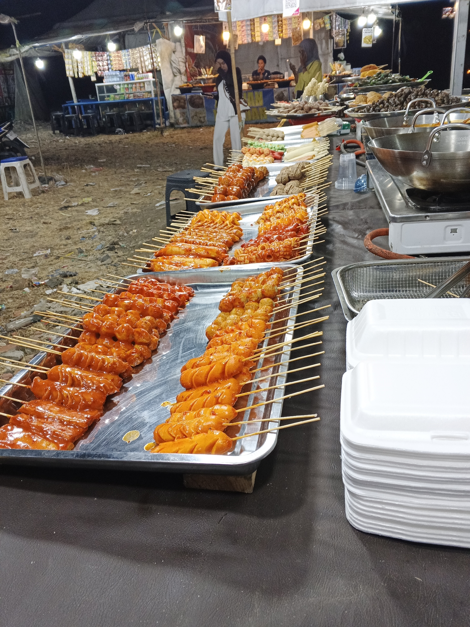 This image depicts an outdoor food stall at night, likely in Sukabumi, Indonesia, displaying a variety of grilled food on skewers. In the foreground, several metal trays are filled with rows of meat skewers, predominantly sausages and what appear to be meatballs, all coated in a reddish-orange sauce. The skewers are arranged neatly on wooden sticks. Behind the food stall, there are tents, lights, and shelves stocked with various food items and drinks, suggesting a bustling night market. In the middle ground, people are visible, some tending to the stall and others browsing. The ground appears to be dirt and debris. To the right of the food stall, a stack of white styrofoam containers is visible, ready for customers. A gas stove with pots is also present, indicating food is being prepared or kept warm. The overall scene suggests a typical street food vendor setup in a market environment at dusk or night.