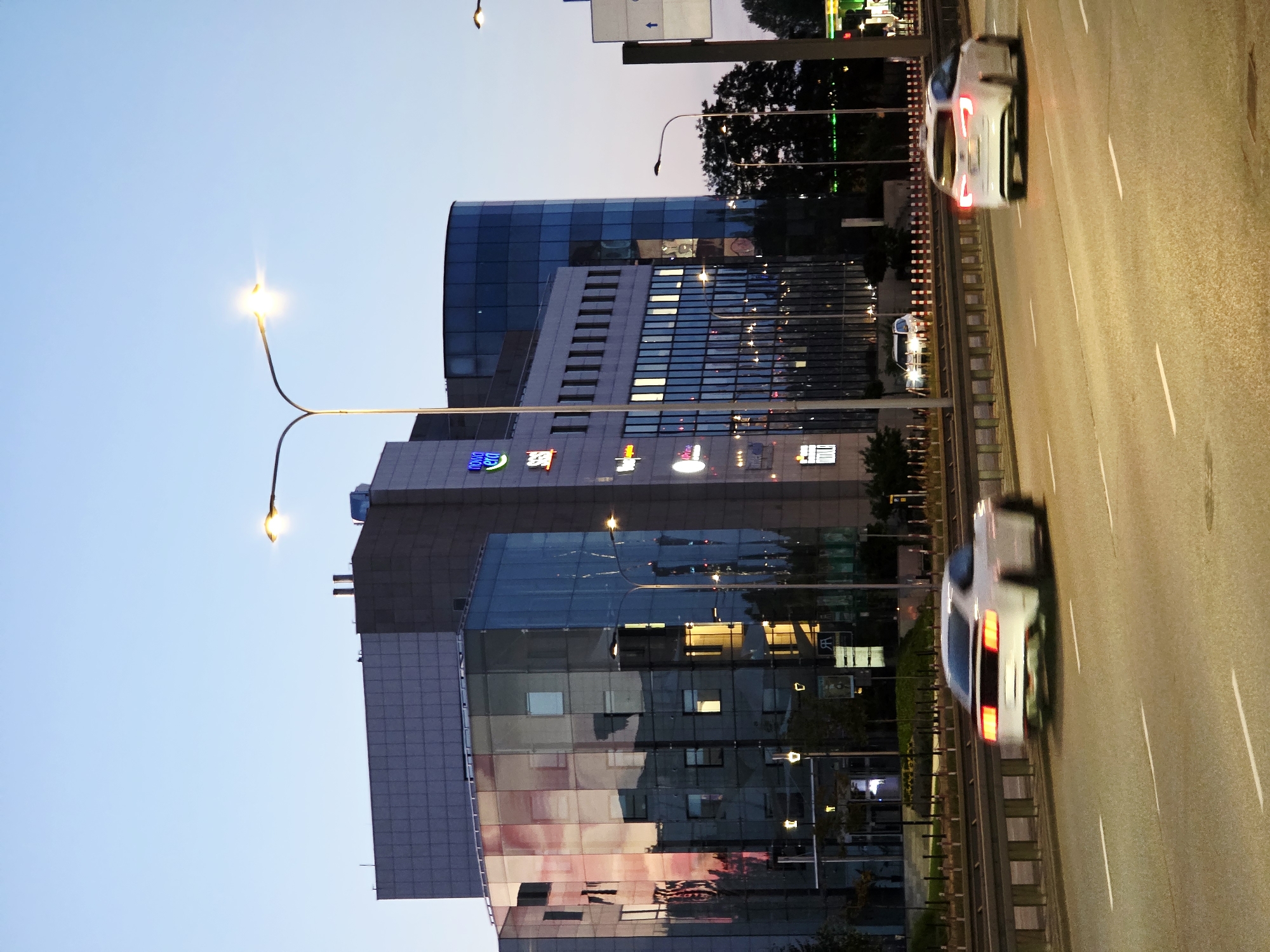 A vertical, eye-level shot shows a street scene at dusk with cars on a highway and modern buildings on the side. The sky is a soft gradient of light blue to lavender, suggesting either early morning or late evening. On the right side of the image, a highway runs diagonally, with two lanes of asphalt visible. White lines mark the lanes, and a metal guardrail separates the highway from the sidewalk. Two cars, one in the foreground and one further down the road, are captured in motion, appearing as streaks of light and blurred shapes. The foreground car is white with its taillights illuminated, indicating it is braking or has its lights on. The background car is also white and appears to be moving away from the camera.
On the left side of the image, a series of modern buildings rise up. The closest building has a facade of dark gray panels and large, reflective windows that mirror the sky and the lights from within. Above this, a larger, curved building features a facade of blue glass windows. The buildings are illuminated from within, with some windows showing bright, warm light, while others are darker. Various signs with colorful logos and text are visible on the lower portion of the buildings, though most are too blurry to read clearly. One sign with blue and white lettering appears to say "NOVO ERA". Streetlights with illuminated bulbs are positioned along the sidewalk. The overall atmosphere is urban and dynamic, with the motion of the cars contrasting with the static architecture.