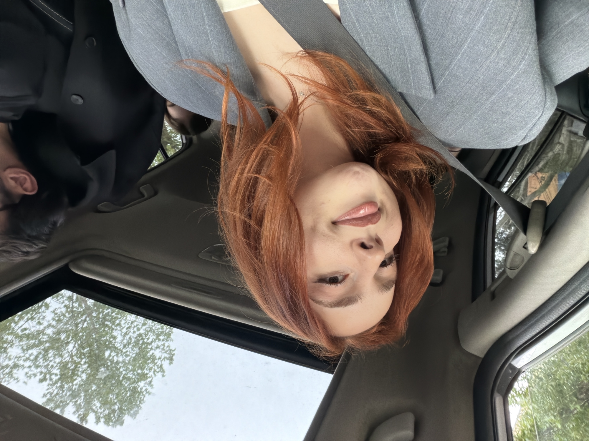 A selfie-style, interior shot taken from inside a car. A woman with reddish-brown hair, wearing a gray blazer, is in the passenger seat, wearing a seatbelt, and appears to be sticking out her tongue slightly. A partial view of a man wearing a black suit is visible in the left side of the frame. The car has a sunroof, through which trees and the sky can be seen. The location cannot be determined as Tbilisi, Georgia.
