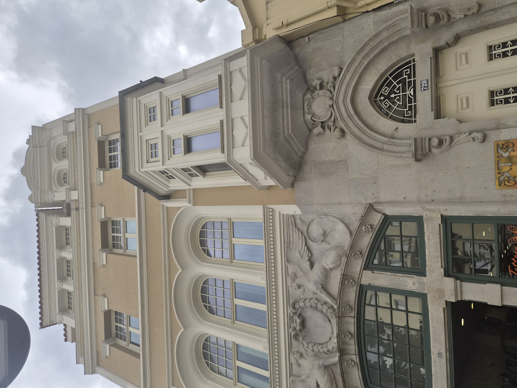 A low-angle, exterior shot captures the ornate facade of a building in Braga, Portugal. The building features a multi-story design with distinct architectural elements. The upper floors are painted a pale yellow and are adorned with decorative white trim, pilasters, and cornices. Several windows with white frames are visible on these levels, some with grid-like panes and others with solid glass.
A prominent bay window, also with white framing, juts out from the middle section of the building. Below this, the facade transitions to a stone material. This section features elaborate relief sculptures depicting winged mythological creatures flanking a central crest. Further down, a large arched doorway with decorative stone carvings is visible, leading to a light-colored double door. Above the doorway, a small window with a geometric metal grille is set into the arch. Two house numbers are visible: "725" above a ground-floor window on the left, and "729" above the arched doorway.
Below the stone relief, a large window with dark green framing is visible on the left side of the building. This window reflects trees and surrounding buildings. To the right of this window, another window of the same style and color is present. A portion of the lower ground floor reveals an open entrance with lighting. A sign with "Tattoo" is partially visible on the left.
The sky above is overcast and filled with gray clouds. The overall impression is one of an older, architecturally rich building in an urban setting.