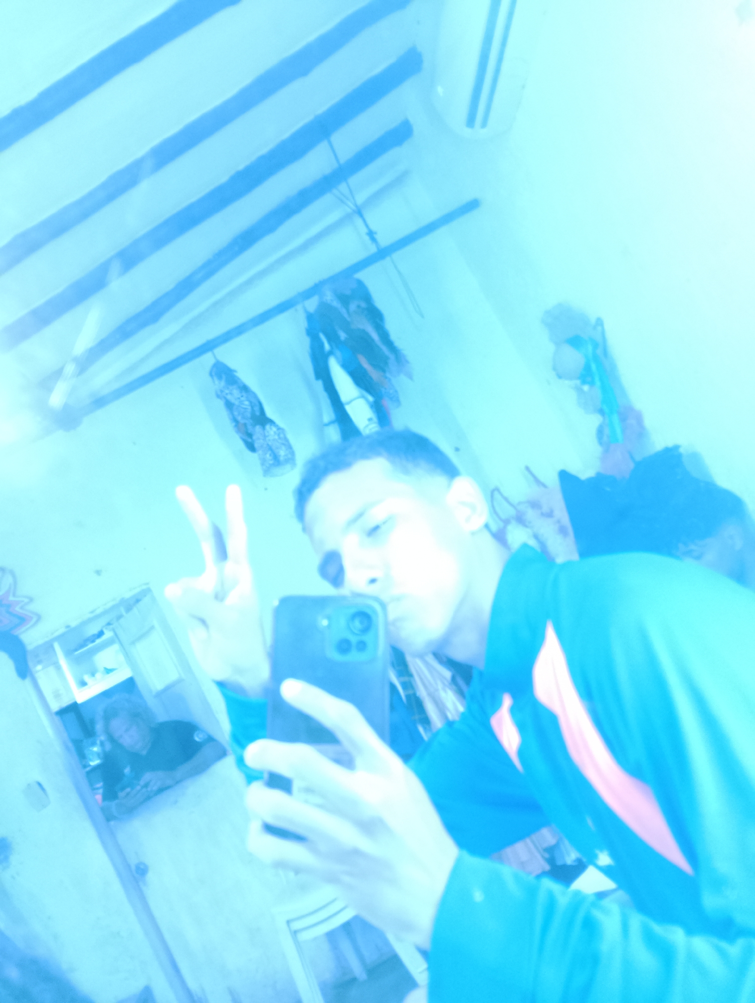 A young man is taking a selfie in a room. The location might be Porlamar, Venezuela. He is holding a smartphone with his right hand and making a peace sign with his left hand. The room appears to be cluttered with items hanging on the wall. In the mirror, there is a reflection of another person using a phone. The room is bathed in blue light. An air conditioner is visible near the top right of the image.