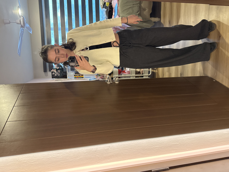 A person poses in a mirror, taking a selfie with their phone. They wear a light yellow cardigan over a black top, paired with dark grey drawstring trousers and dark shoes. The background includes a window with blinds, a ceiling fan, and various items on a shelf. The mirror reflects a wooden door and a neutral-toned wall with a light strip at the bottom. The image is rotated.
