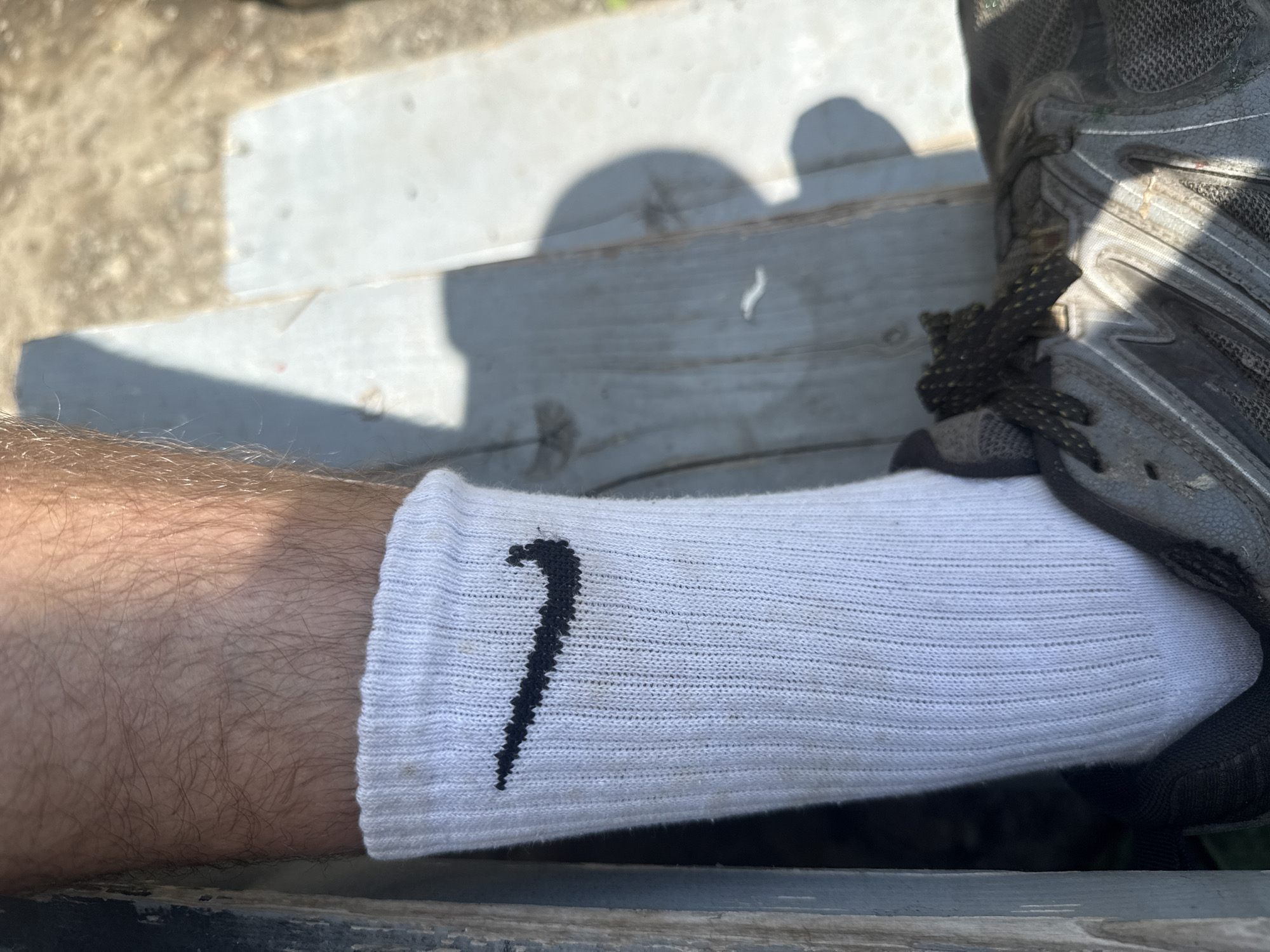 A close-up, overhead shot shows a person's left leg and foot, wearing a white Nike sock with a black Nike logo and a gray athletic shoe. The leg is hairy, and the sock is pulled up to mid-calf. The shoe laces are dark and textured. The background is a wooden surface, possibly a bench or deck, in an outdoor setting. Sunlight casts shadows across the scene. The overall impression is casual and outdoor-oriented.