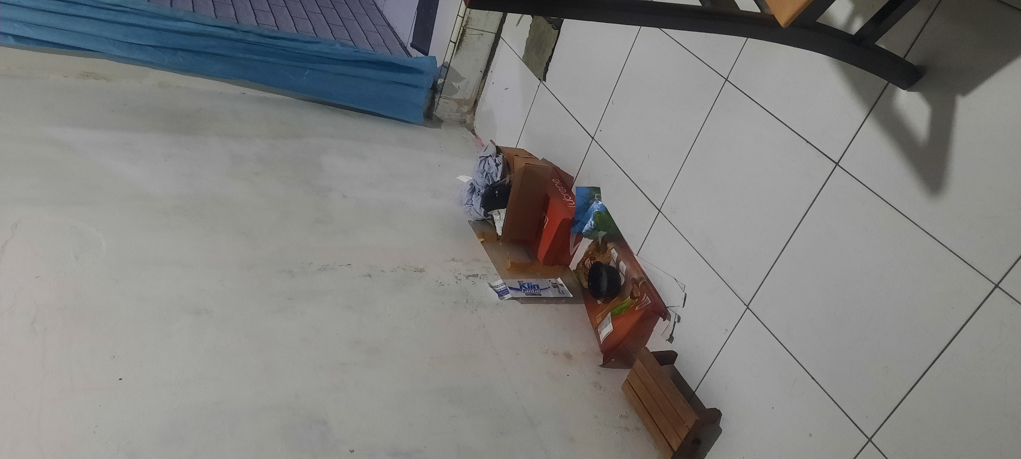 The image depicts an indoor scene, likely a corner of a room. The walls are a light, off-white color with some visible staining and marks, suggesting age or environmental factors. The floor is tiled with large, square white tiles, with dark grout lines creating a grid pattern.
In the foreground, a small, simple wooden stool with a slatted seat is placed on the floor to the left. Adjacent to it, a low, flat object, possibly a large cardboard box or mat, is laid out. On this object, there is a small black bowl.
Further to the right, a stack of cardboard boxes is visible. One prominent box is red and appears to be a container for "Lubrene." Above and behind this, other cardboard boxes are present, some containing crumpled clothing or fabric. A blue curtain is visible on the far right, hanging against what appears to be a window or doorway with a brick-like pattern visible behind it.
The overall impression is of a cluttered or storage area, possibly a storage room or a corner of a living space where items are temporarily placed. The lighting suggests it might be evening or a dimly lit interior.
There are no people visible in the image. The scene is static and appears to be a still life capture of a corner of a room.
There is no visual information in the image that would allow for a specific location within the city of North Jakarta, Indonesia. The scene is generic and could be found in many locations.