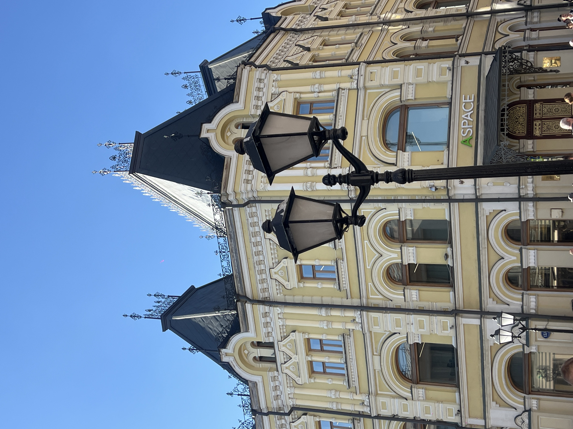 This is a medium shot, eye-level, outdoor photograph of a historical building in Moscow, Russia. The building is painted a pale yellow with white decorative trim and features a dark grey, mansard-style roof with ornate metal cresting. Several windows with arched tops and brown frames are visible on the facade. In the foreground, a black, wrought-iron street lamp with two lantern heads is positioned on the right side of the frame. Below the lamp, a green and white sign reads "A SPACE". The sky is a clear, bright blue. The image appears to have been taken on a sunny day.