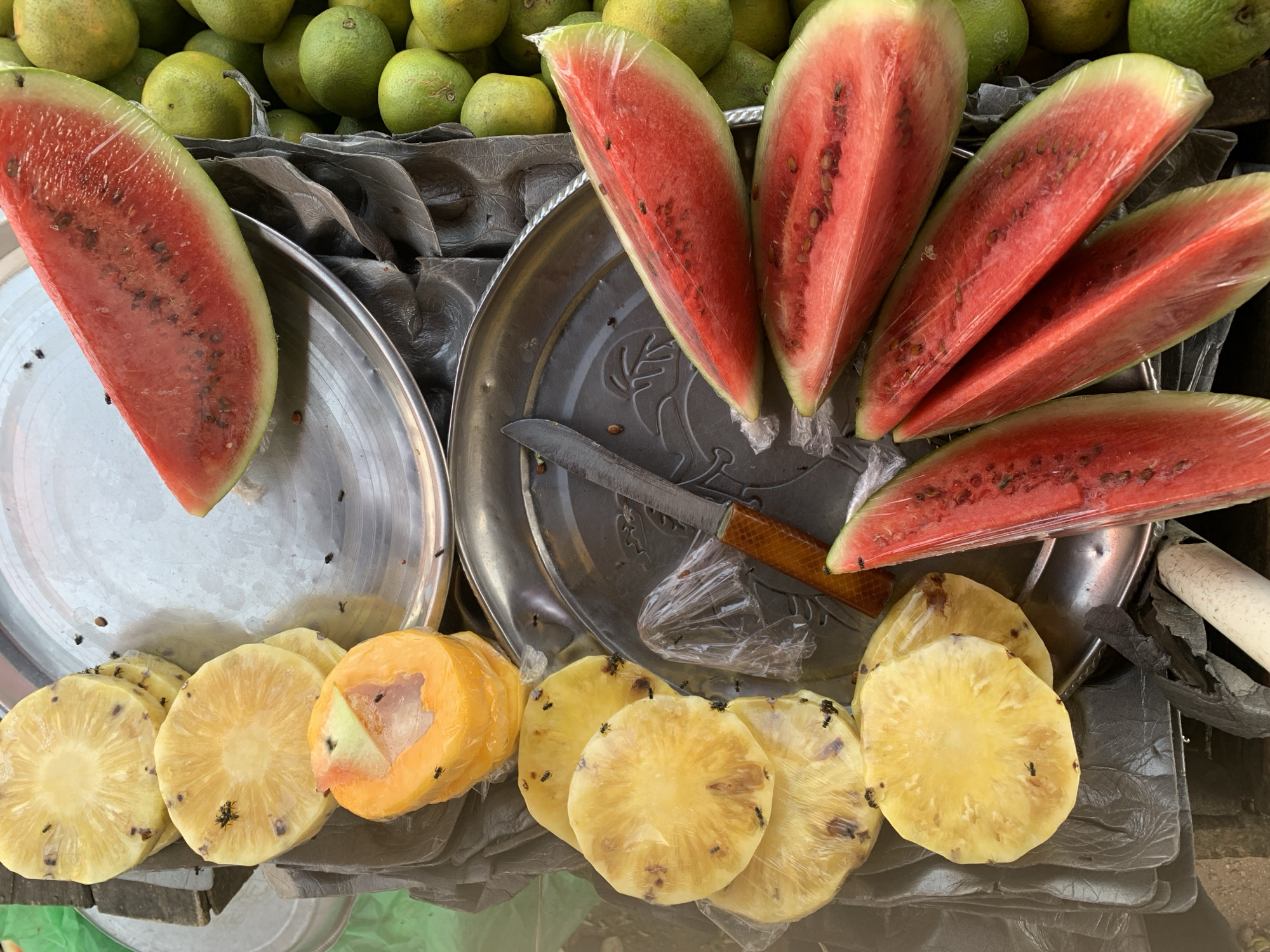 The image shows a fruit stand, likely in a public market. The prominent items are watermelon slices wrapped in plastic, pineapple slices, and unidentified green fruits. These items sit on metal trays. A knife with a brown handle is visible on one of the trays. Flies are present on the pineapple and other fruits. The presence of fruits cut and displayed indicates a vendor selling prepared fruit. The location is not determinable from visual information alone. The city is implied to be New Stadium, Nigeria, by the prompt.