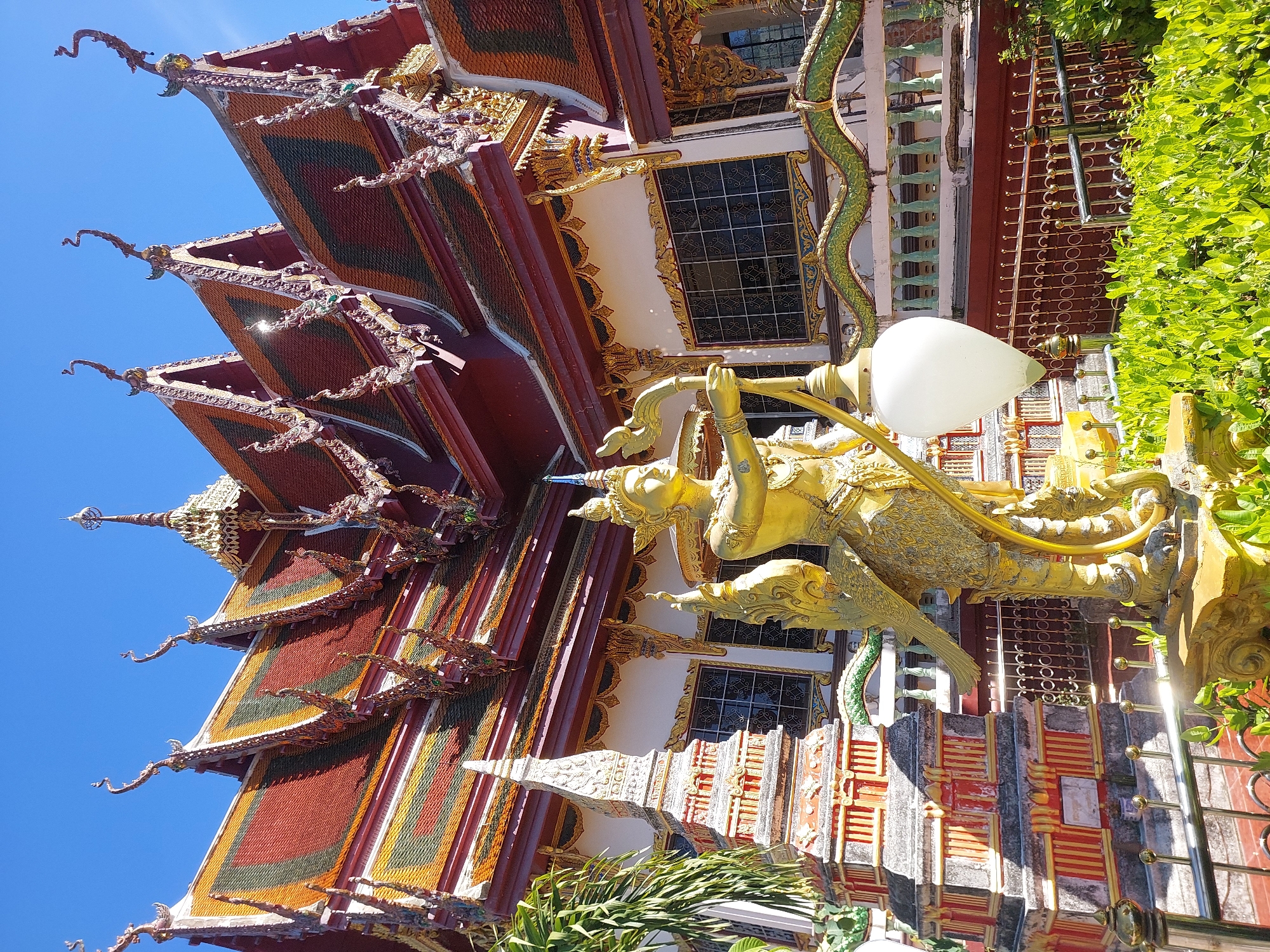 This media file captures a vibrant, ornate temple structure, likely a Buddhist temple, with a clear blue sky overhead, suggesting a sunny day. The architecture features multiple tiers of steeply pitched roofs, adorned with colorful tiles in shades of red, yellow, and green. Decorative elements, including what appear to be mythical creature sculptures and intricate carvings, are visible along the rooflines and eaves.
In the foreground, a striking golden statue of a mythical being with bird-like features and human-like arms is prominently displayed. The statue is holding an object resembling a horn or flute and is intricately detailed. Beside it, a large, white, lotus-shaped lamp or light fixture adds to the decorative richness. A coiled, green, serpent-like sculpture also winds its way up the side of the building, adding to the mythological imagery.
The building itself exhibits a facade with decorative windows framed in gold and patterns of red and white. A metal fence with circular designs and a red wall are also visible. Lush green foliage partially frames the scene on the right, contrasting with the bright colors and gold of the temple.
The overall impression is one of a spiritual and culturally rich setting, possibly a tourist attraction given the well-maintained and elaborately decorated nature of the temple. The sunlight casting reflections indicates it is daytime. The scene evokes a sense of awe and wonder due to the intricate artistry and the symbolic imagery. No visible text is present in the media file. The location context provided is Baan Plai Laem, Thailand, which aligns with the architectural style and cultural motifs depicted.