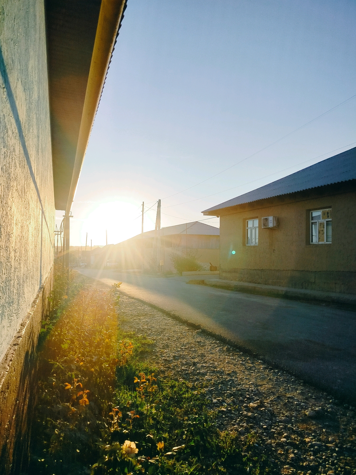 The image depicts a street scene at sunset in the city of Kölkent, Kazakhstan. The sun is low on the horizon, casting long shadows and creating lens flare across the road. On the left, a textured, light-colored wall with a yellow overhang occupies the foreground. To the right of the wall, a patch of green grass and some yellow flowers grow along the edge of a gravel and asphalt road. In the mid-ground, a simple, light brown house with a dark, corrugated roof is visible. The house has two windows and an air conditioning unit mounted on its exterior wall. The road stretches into the distance, lined with utility poles. The sky is clear and a pale blue, with the sun's rays creating a bright, hazy atmosphere. No people are visible in the frame.
