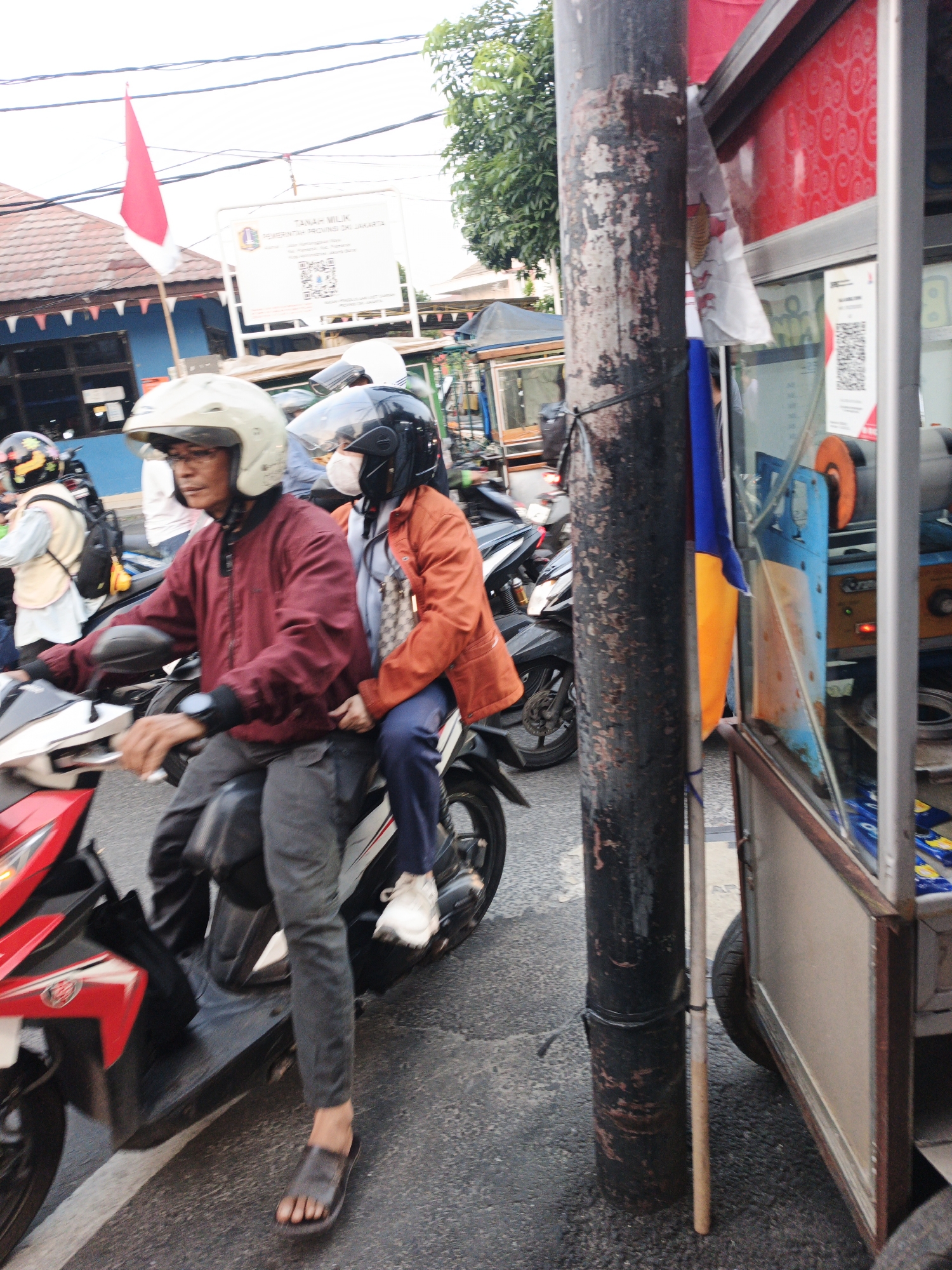 Two individuals are riding on a motorcycle on a street in West Jakarta, Indonesia. The driver, a male wearing a red jacket and a white helmet, is looking forward. The passenger, a female wearing an orange jacket and a black helmet, is partially obscured. The motorcycle is red and black. In the background, there is a street vendor's cart with red and white accents, a utility pole, and a sign that appears to be in Indonesian. The street is paved with asphalt. The scene is likely captured during daylight.