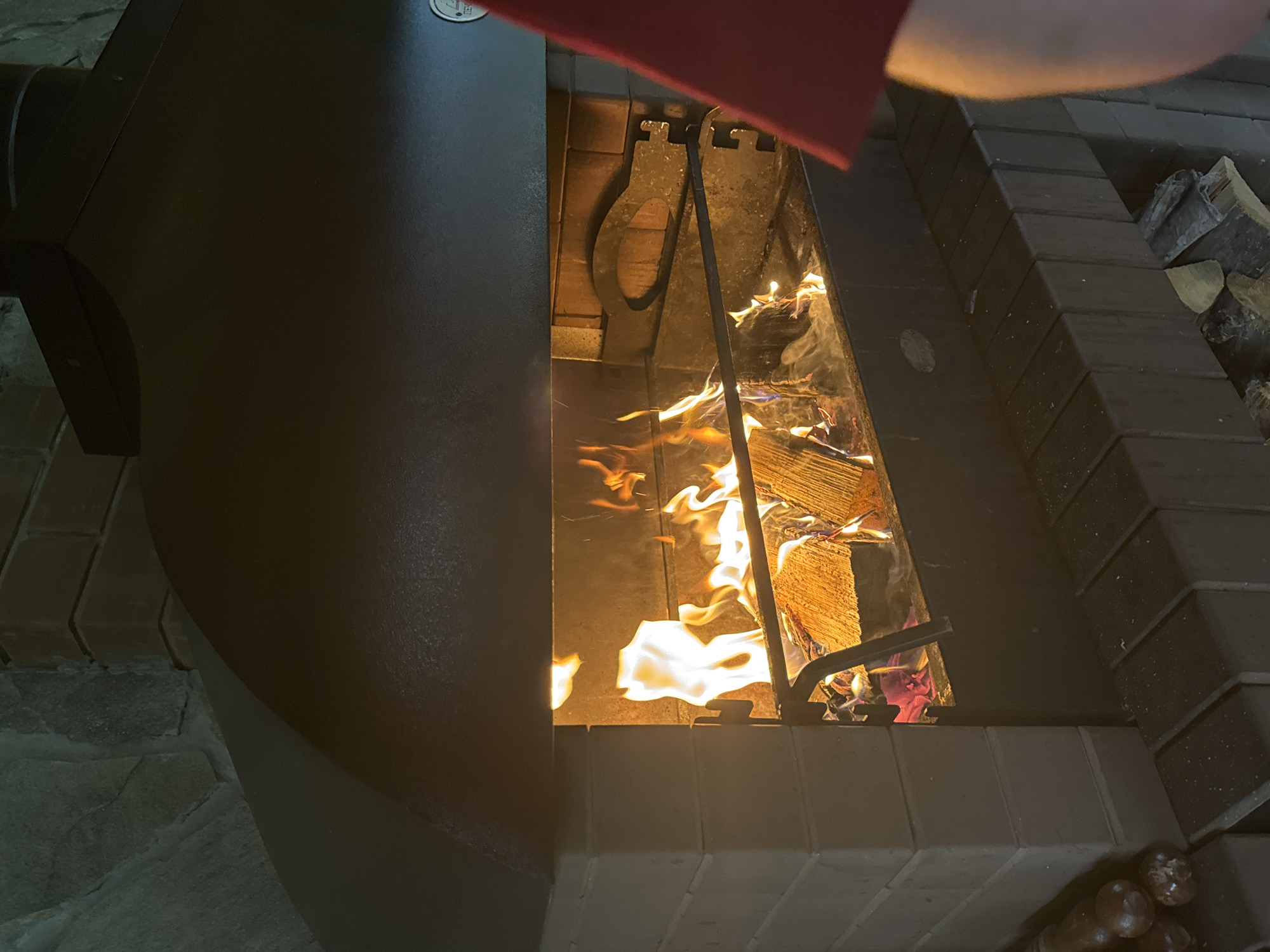 An indoor fireplace is filled with burning logs. The fireplace is constructed of light-colored bricks and has a dark metal hood. A person wearing a red shirt is partially visible on the right side of the image. Wood is stacked in the lower portion of the fireplace structure. The scene appears to be from a home in Ryabinino, Russia.