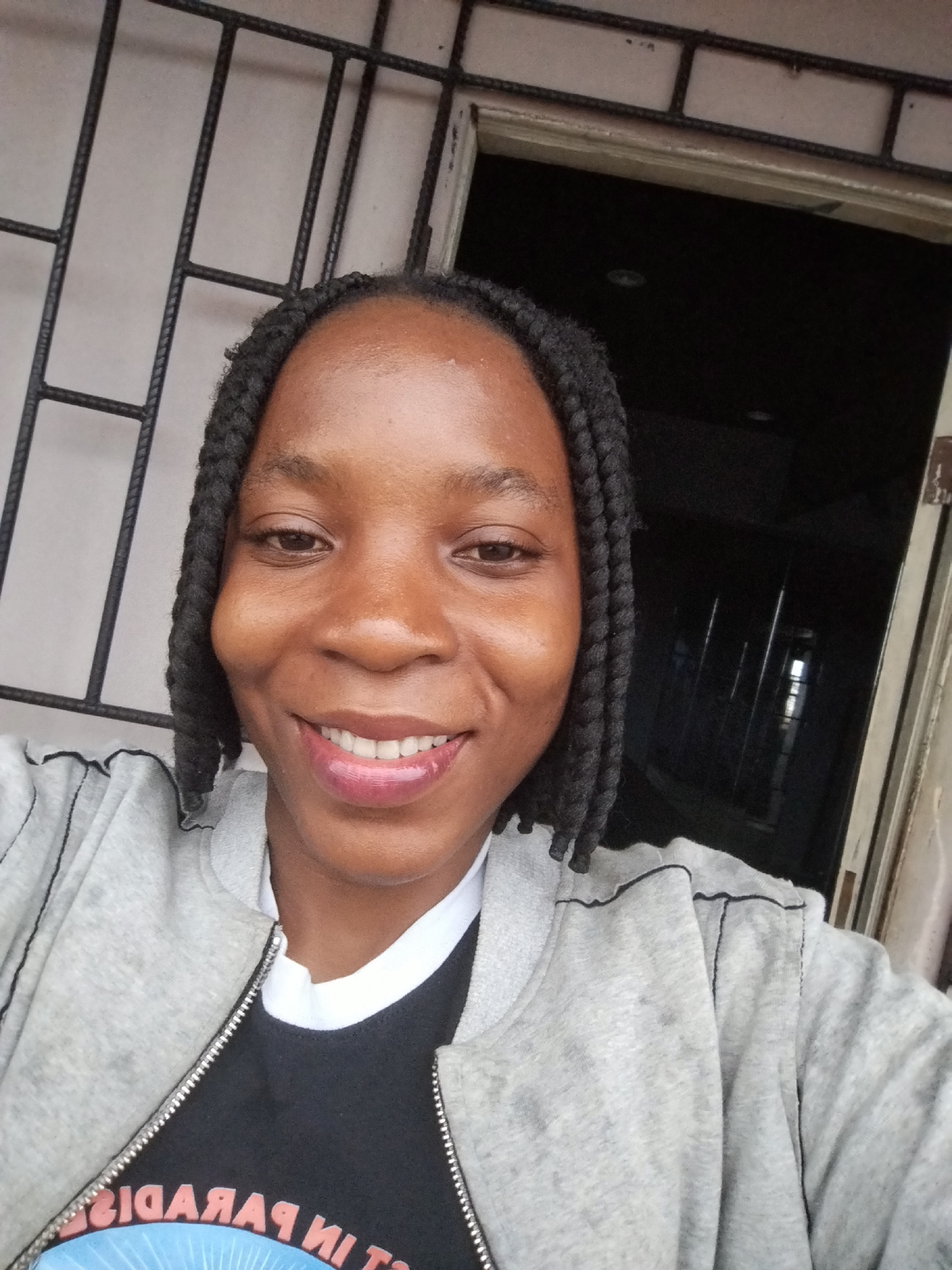 A young Black woman with dark braided hair pulled into two thick braids extending past her shoulders, smiles at the camera. She is wearing a light grey zip-up jacket over a black t-shirt with red text. The image appears to be taken outside a building, with a barred window visible in the background to the left and a dark doorway or opening to the right. The overall setting suggests a casual, outdoor urban environment, possibly in Uselu, Nigeria.