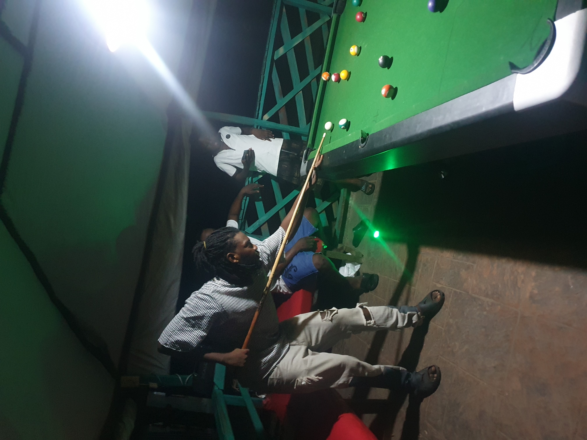 In Lokoja, Nigeria, a group of young Black men are playing pool outdoors at night. The game is taking place on a green felt pool table under the cover of a canvas awning, illuminated by green and white lights.
The man in the foreground, wearing a striped shirt and ripped light-colored pants, is in the process of striking a pool ball with a cue stick. He is facing the table and appears focused on his shot. To his right, another man in a white t-shirt and dark shorts is observing the game. Behind them, another individual in a white shirt is visible, holding a mobile phone.
The pool table is set up on a textured, reddish-brown tiled floor. The environment appears to be an open-air space, with a railing and posts visible in the background, suggesting it might be a patio or a dedicated recreational area. Several colored pool balls are scattered across the table. The lighting creates a vibrant atmosphere, with green light casting a glow on the table and surrounding areas.