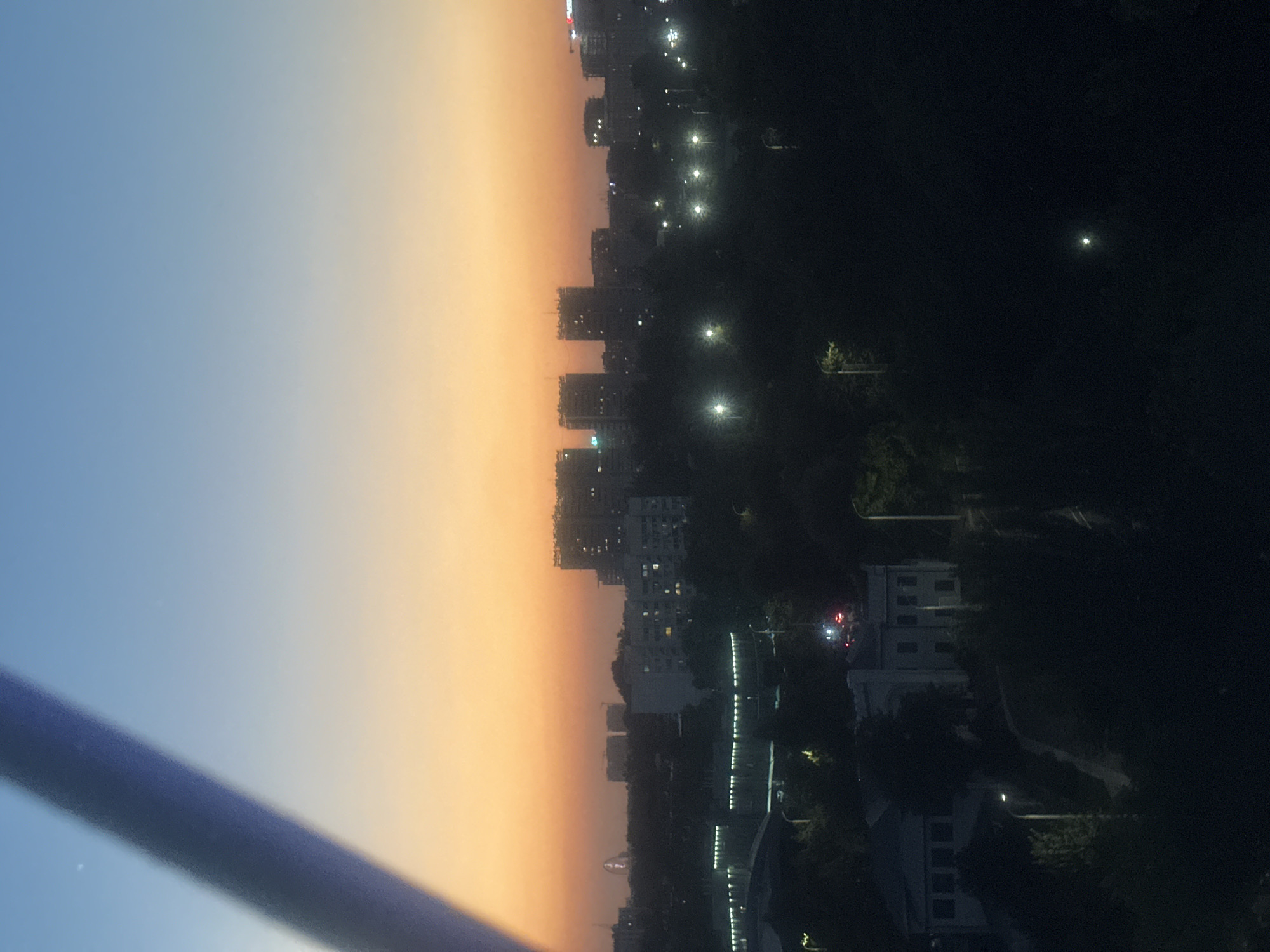 The image shows an urban landscape at dusk, with buildings silhouetted against a gradient sky transitioning from pale blue to orange. A diagonal dark object, possibly a railing or a structural element of a balcony, traverses the lower left portion of the frame. In the distance, the city lights are beginning to twinkle, indicating the onset of night. The scene is characterized by the dimming light and the urban development, suggesting a residential or commercial area. The time of day is clearly dusk, with the sky exhibiting the characteristic colors of sunset or sunrise. The location is identified as Tashkent, Uzbekistan, which would imply the architectural style of the buildings and the overall urban planning are consistent with that region. No specific activities or events are discernible, and no people are visible. The overall mood is serene and contemplative, evoked by the transition from day to night over a cityscape.