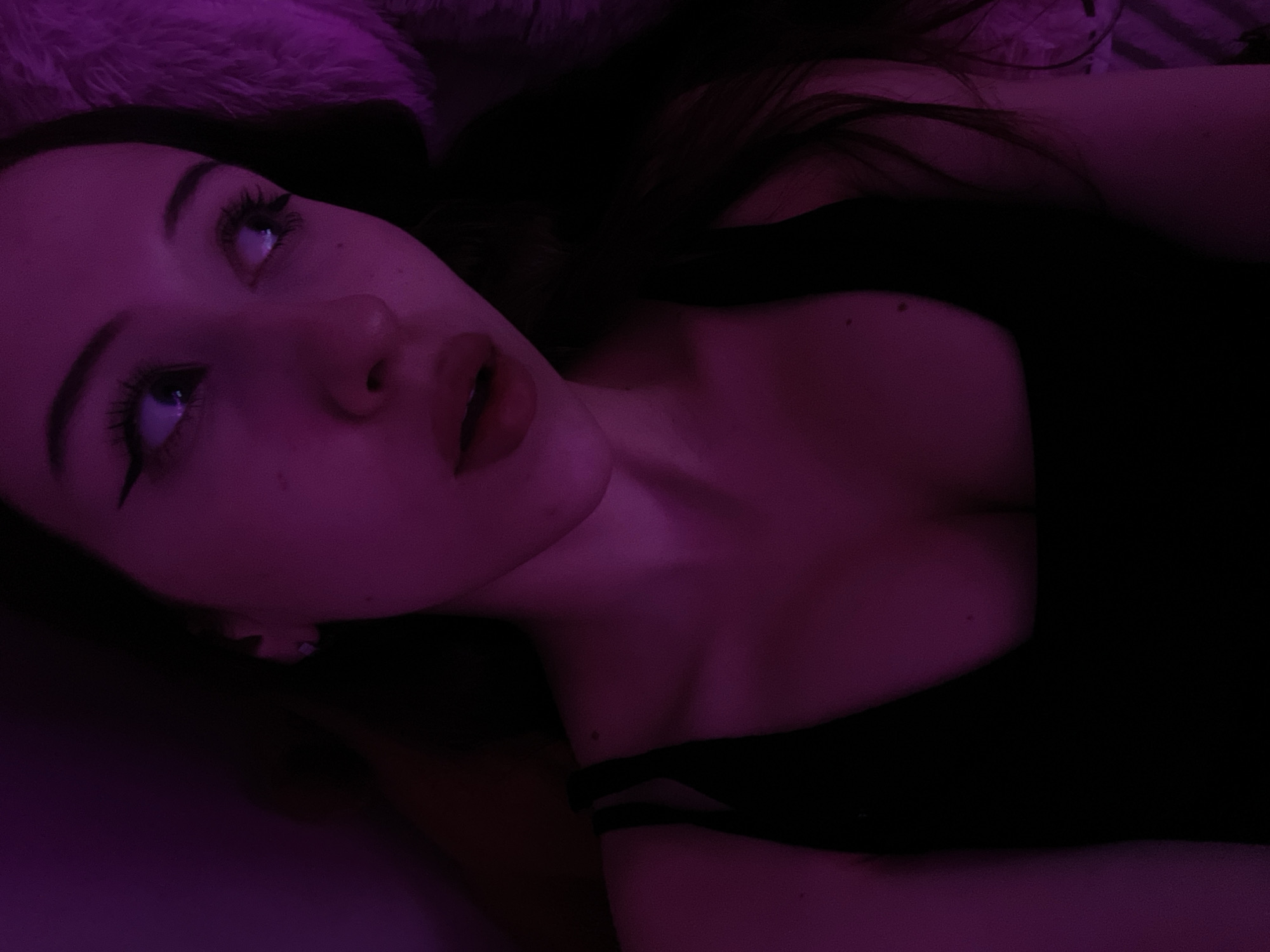A young woman is depicted in a close-up, low-light portrait, illuminated by a vibrant purple hue. She is wearing a black, low-cut top, and her face is angled slightly away from the camera, with her gaze directed to the right. Her eyes are accentuated with winged eyeliner, and her lips are parted. The background is dark, with a hint of a fluffy, textured surface visible to her right. The image does not contain any identifiable landmarks or context that would allow for the determination of its location in Votkinsk, Russia.