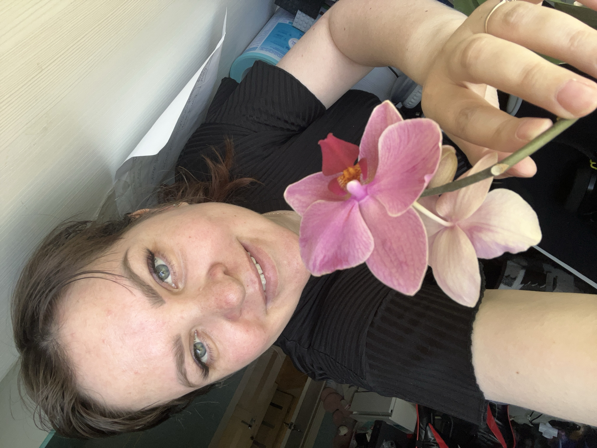 A woman with fair skin and short brown hair is holding a stem with two pink orchid flowers in her right hand. She is wearing a black ribbed shirt. A silver ring is visible on her right ring finger. In the background, there is a white wall, a piece of paper with writing, and a blue bottle. No indication of the location being in Noviy, Russia is visible.