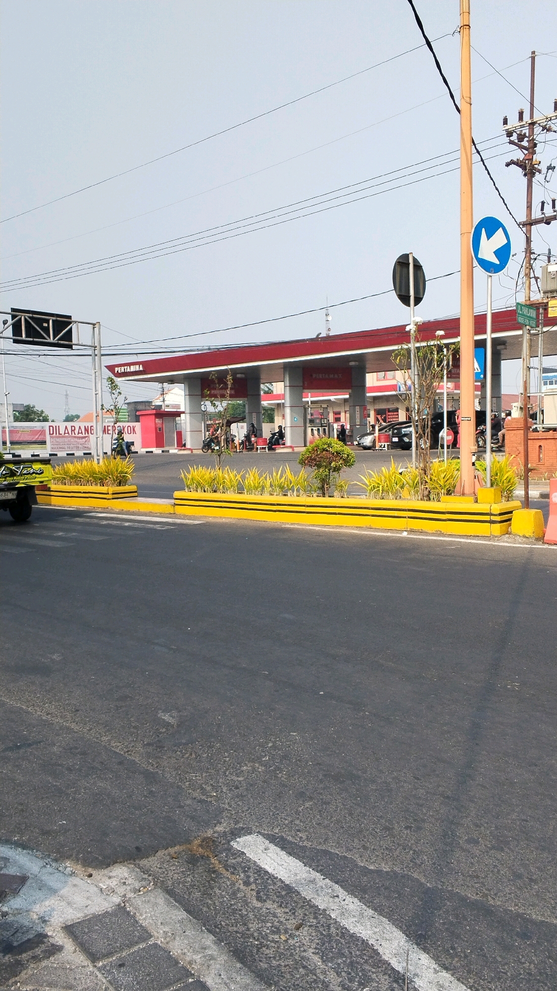 The image shows a Pertamina gas station in Mojokerto, Indonesia, on a bright, clear day. In the foreground, a paved road with white crossing lines is visible. A yellow planter box with black stripes and yellow-green plants runs along the edge of the road, separating it from the gas station. Behind the planter box, a Pertamina gas station with a red roof and white pillars is the dominant structure. Several cars and motorcycles are parked or in motion at the gas station. A sign with red lettering reading "DILARANG MEROKOK" (No Smoking) is visible on the left side of the image. Electrical poles with wires are present on the right side of the frame. The sky is clear blue with some faint power lines stretching across it. The overall scene depicts a typical urban environment with commercial activity.
