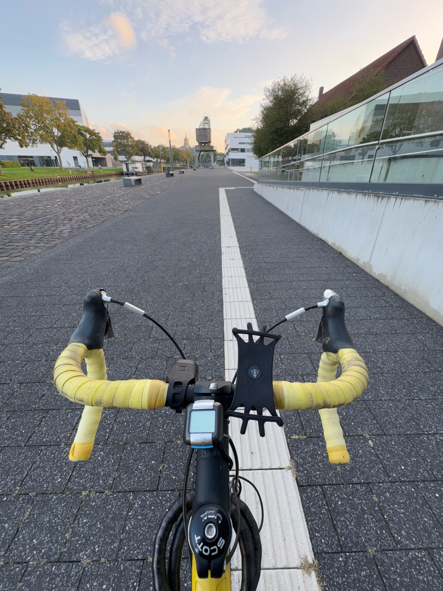 The image is a first-person perspective from a bicycle handlebar, showing a paved path with a white stripe marking the center, leading towards a cityscape in the distance. The path is bordered by a channel of water on the left and a modern building with a glass facade on the right. The bicycle's yellow handlebar tape is visible, along with a phone mount and a cycling computer. The sky is partly cloudy with the soft light of early morning or late afternoon. The cityscape includes buildings of varying architectural styles and a prominent industrial crane. The location is the Rhine-Waal University of Applied Sciences campus in Kleve, Germany, specifically along the Rhine riverfront. There are no people or animals visible in the frame.