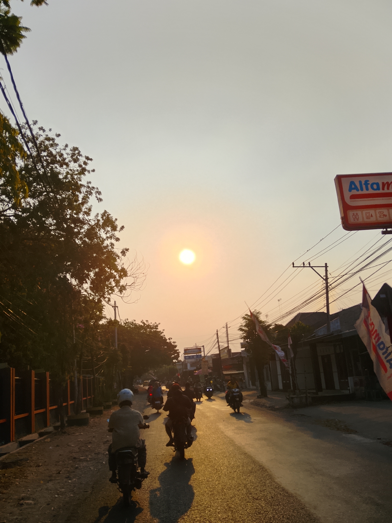 The image depicts a street scene in Ngelo, Indonesia, during sunset. The sky is a pale orange, with the sun appearing as a bright, diffused orb in the upper center. A road runs diagonally from the bottom left to the upper right, reflecting the warm light of the setting sun. Several people on motorcycles are visible, traveling in both directions. In the foreground, two individuals on motorcycles are heading away from the viewer. To the left, trees line the side of the road, casting long shadows. On the right side of the road, buildings and a sign for "Alfamar" are present. Numerous power lines crisscross the sky. The overall atmosphere suggests a typical evening commute in a semi-urban or rural Indonesian setting.