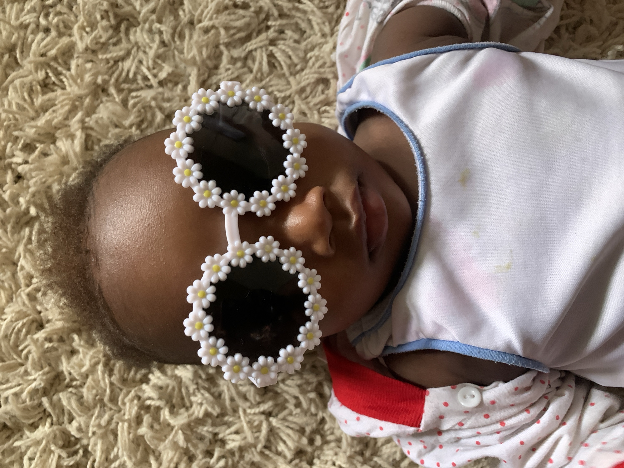 An infant lies on a beige rug, wearing sunglasses adorned with white daisy-shaped frames. The infant wears a white garment with a blue trim and a red and white polka-dot sleeved garment underneath. The scene's location in Barakin Akawo, Nigeria is undetermined.