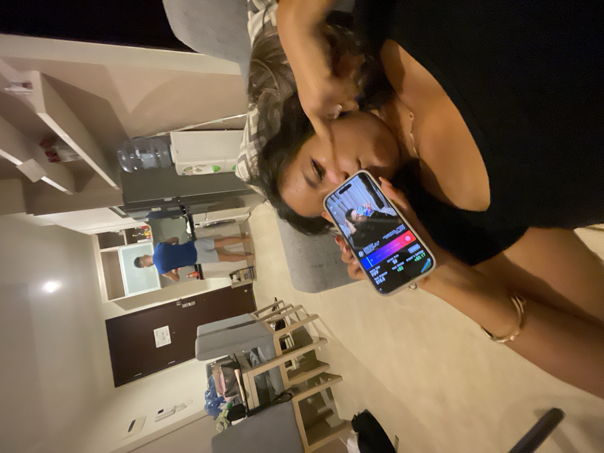 The image shows a woman holding a smartphone, displaying a game or app interface. In the background, a man in a blue shirt and gray shorts is standing in what appears to be a kitchen area. The location seems to be indoors. It can be inferred from the EXIF metadata of the picture that the image was captured in Central Jakarta, Indonesia.
