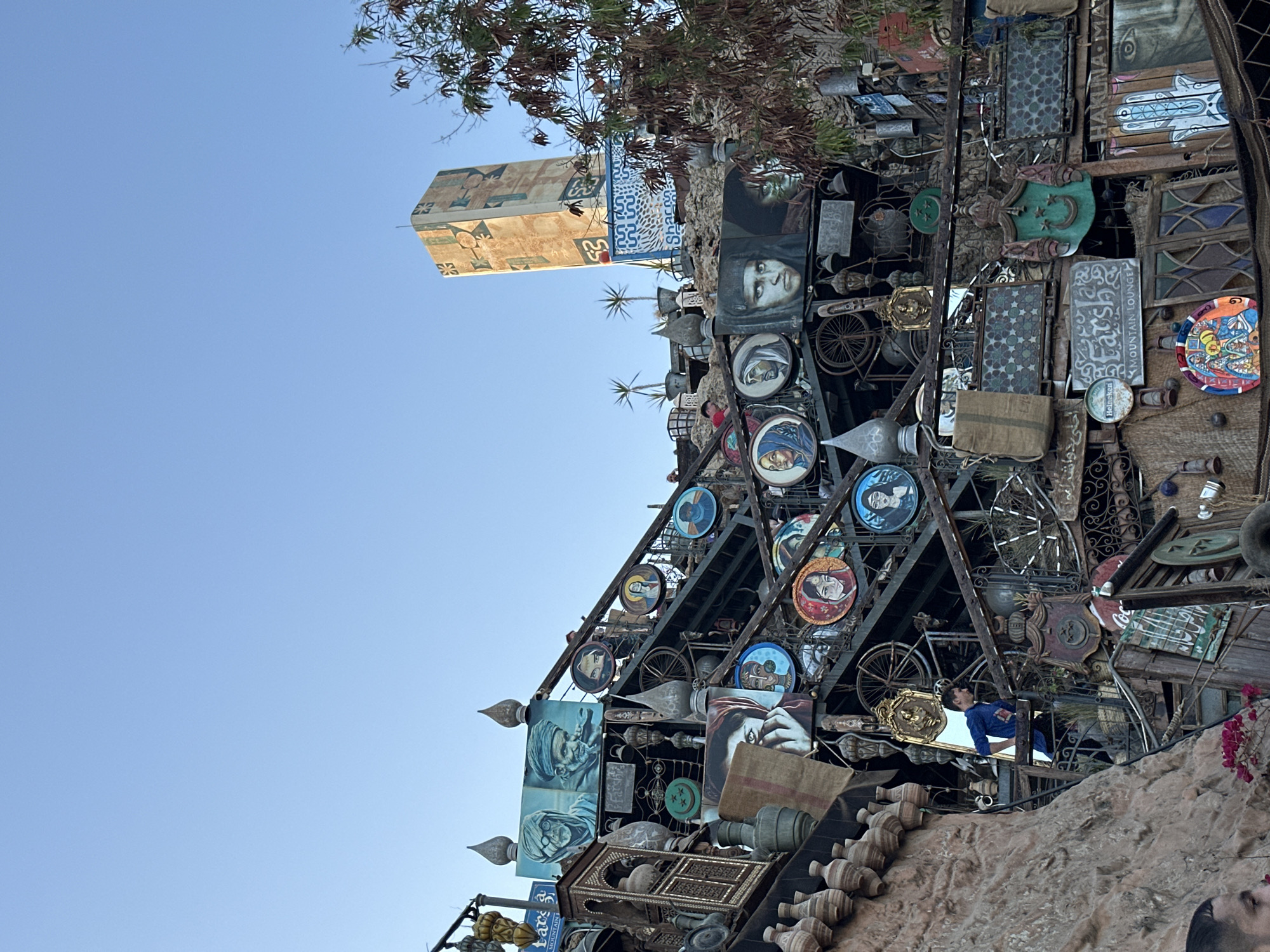 This image captures a vibrant and eclectic scene, likely in Sharm El Sheikh, Egypt, showcasing a large, multi-tiered structure or building facade heavily adorned with various decorative objects. The main subject is this elaborate installation, which features a complex framework made of metal, possibly wood, layered with an array of artistic and functional items.
Prominently displayed are numerous paintings, many of which are portraits of people, some appearing in traditional Middle Eastern attire. These portraits are often set within circular frames or on square canvases. Interspersed among the paintings are decorative metal plates, bicycle wheels, lanterns, and various metallic and wooden ornaments. Other items include burlap sacks, pottery, and small mirrors, one of which reflects a man in a blue shirt. The structure also incorporates stained glass or patterned panels with geometric designs.
In the background, a tall, light-colored building with intricate geometric patterns can be seen against a clear blue sky, suggesting a bright daytime setting. Below some dark green foliage, a blue sign with Arabic script and the word "SHARQ" is visible next to this building. The foreground consists of rough, rocky earth, dotted with some pink flowering plants.
Visible text includes "Farsh Mountain Lounge" on a white sign, "Heineken" on a circular coaster-like item, and partially visible "HAKUNA MAT..." which is likely "Hakuna Matata." The overall aesthetic is rich in cultural detail, blending art, craft, and repurposed items to create a unique and visually engaging display. No specific activity is depicted beyond the static arrangement of these items, creating a detailed and ornate visual spectacle.