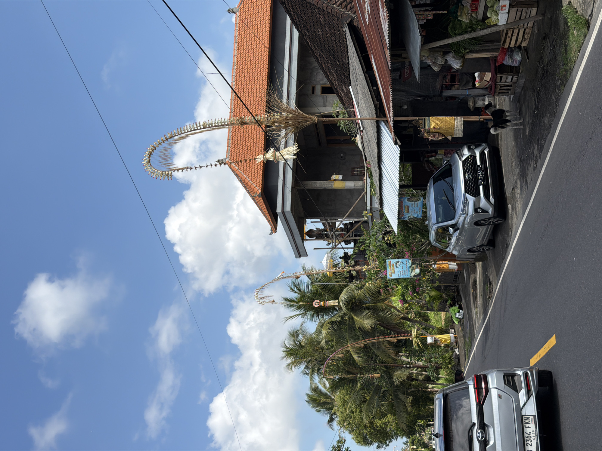 The image depicts a street scene in Tampaksiring, Indonesia, under a partly cloudy blue sky. On the right side of the road, a small shop displays various goods, including produce in wooden crates and bags of items hanging under a weathered metal roof. A black and white dog stands near the shop. In front of the shop, a silver sedan is parked. Further down the street, palm trees and tropical foliage line the roadside, with several tall, decorative bamboo poles adorned with offerings visible. A building with an orange tiled roof and a gray facade is under construction in the background. Two cars are visible on the paved road, one silver sedan in the foreground facing away from the camera, and another silver sedan parked on the right side. A yellow line marks the edge of the road. Electrical wires are strung across the upper portion of the frame.