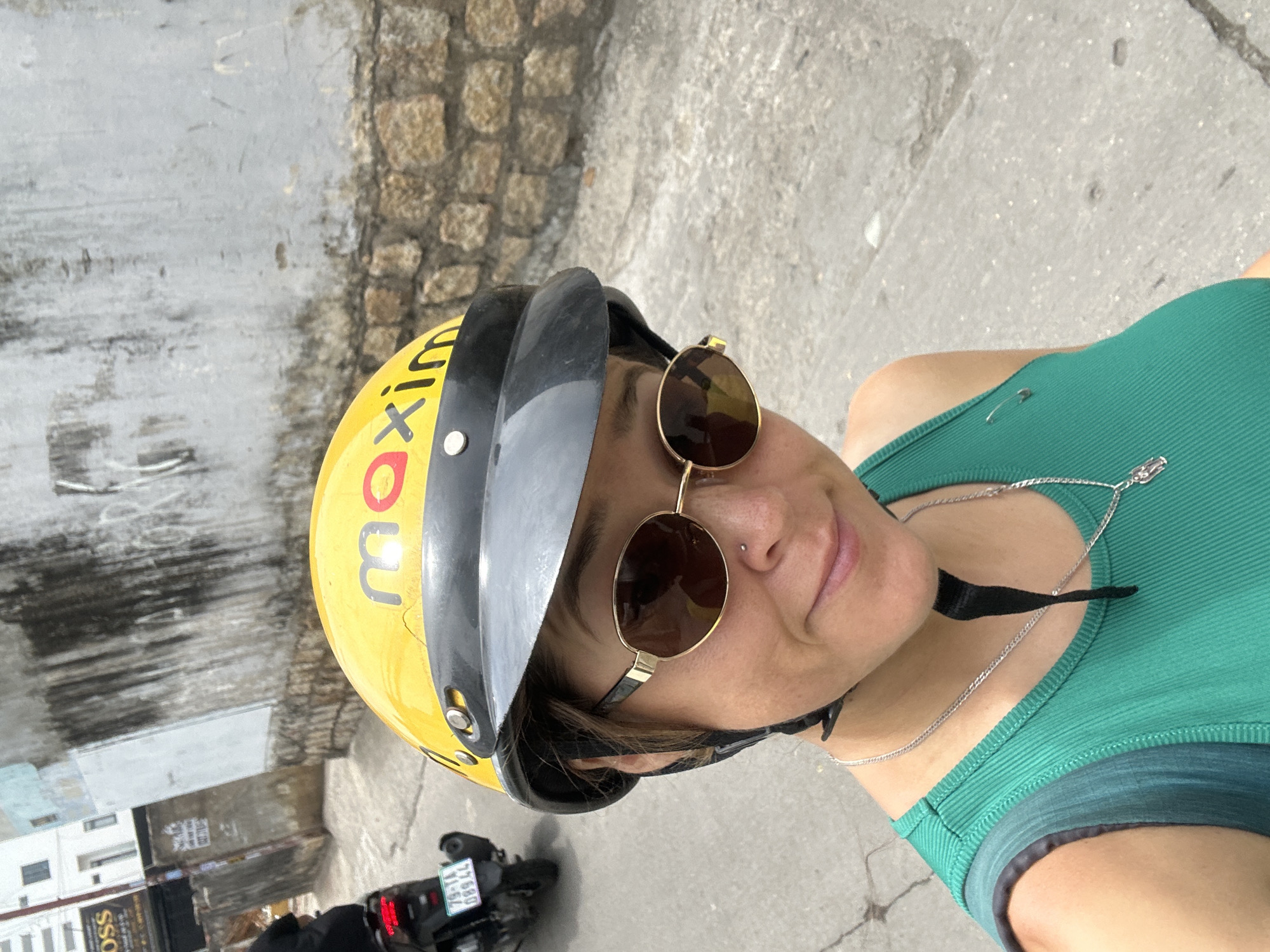 A woman in a green tank top, wearing sunglasses and a yellow "maxim" helmet with a black visor, is positioned in front of a concrete street and wall composed of stones. Part of a motorbike, with a visible license plate reading "79-T1 08944", is behind her. The image was taken in Bac Nha Trang Ward, Vietnam.