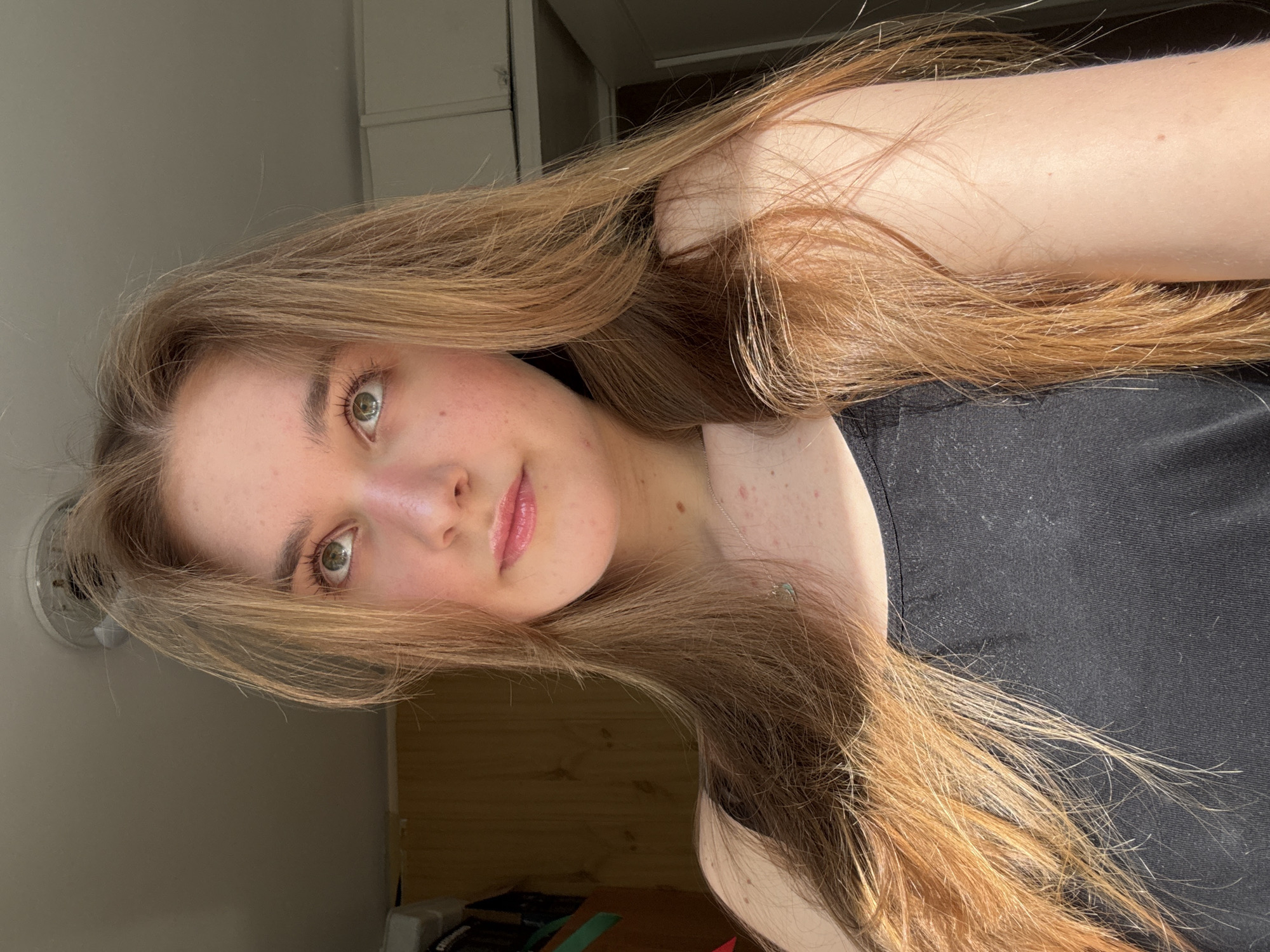 A young woman with long, light brown hair is posing for a selfie. Her hair is partially draped over her right shoulder and arm. She is wearing a black tank top. The background includes a pale wall with a vent and a wooden cabinet or closet. The location cannot be determined as Perm, Russia without more information.