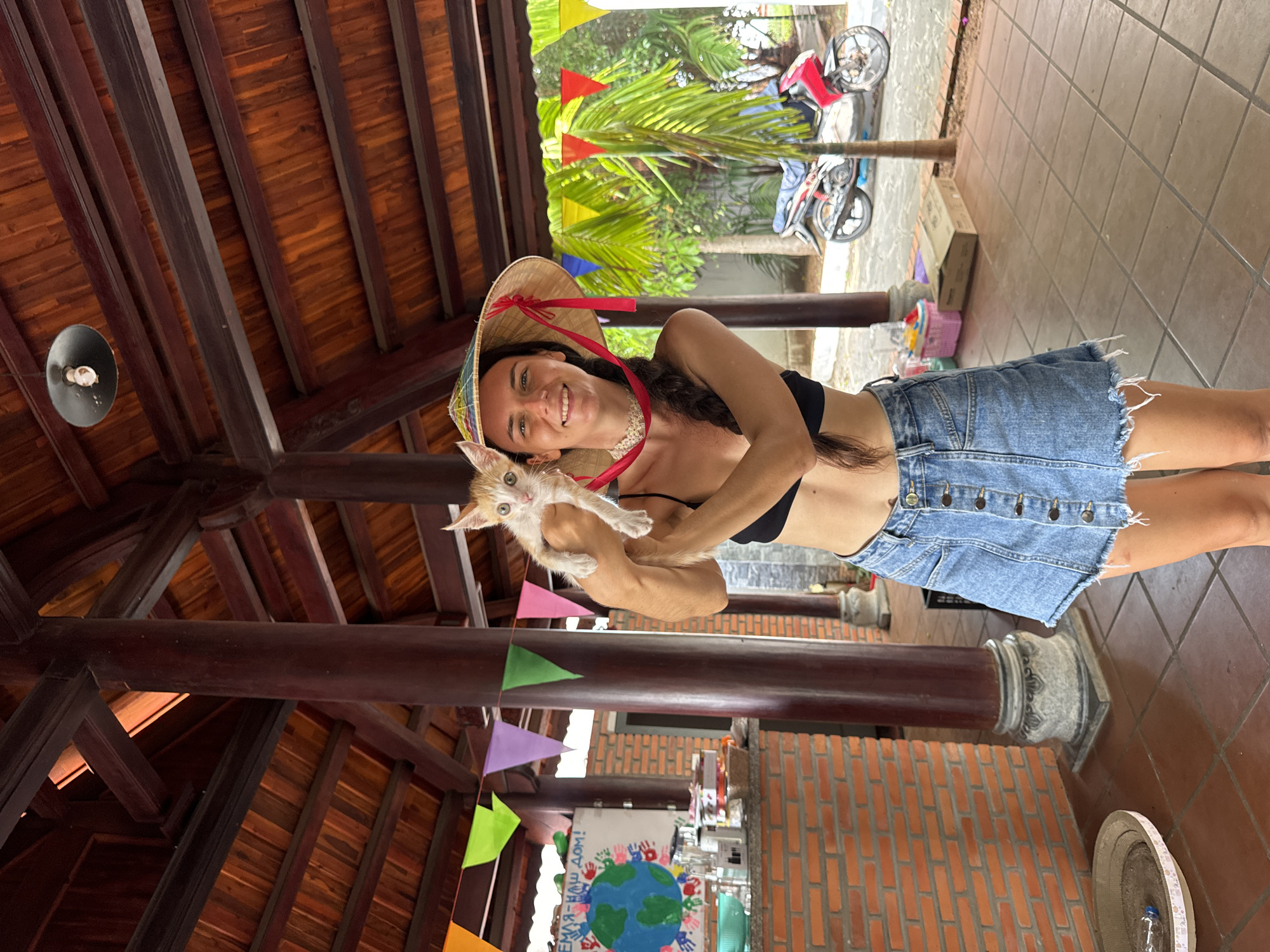 A woman wearing a black crop top, a denim skirt, and a conical hat stands holding a small, light-colored kitten. She is smiling at the camera. Behind her, a red and white motorcycle is parked. The scene appears to be outdoors, under a wooden-roofed structure, possibly a market or cafe, in Mũi Né, Vietnam. The background includes palm trees and brick walls. The ground is tiled. Flags are strung across the scene.