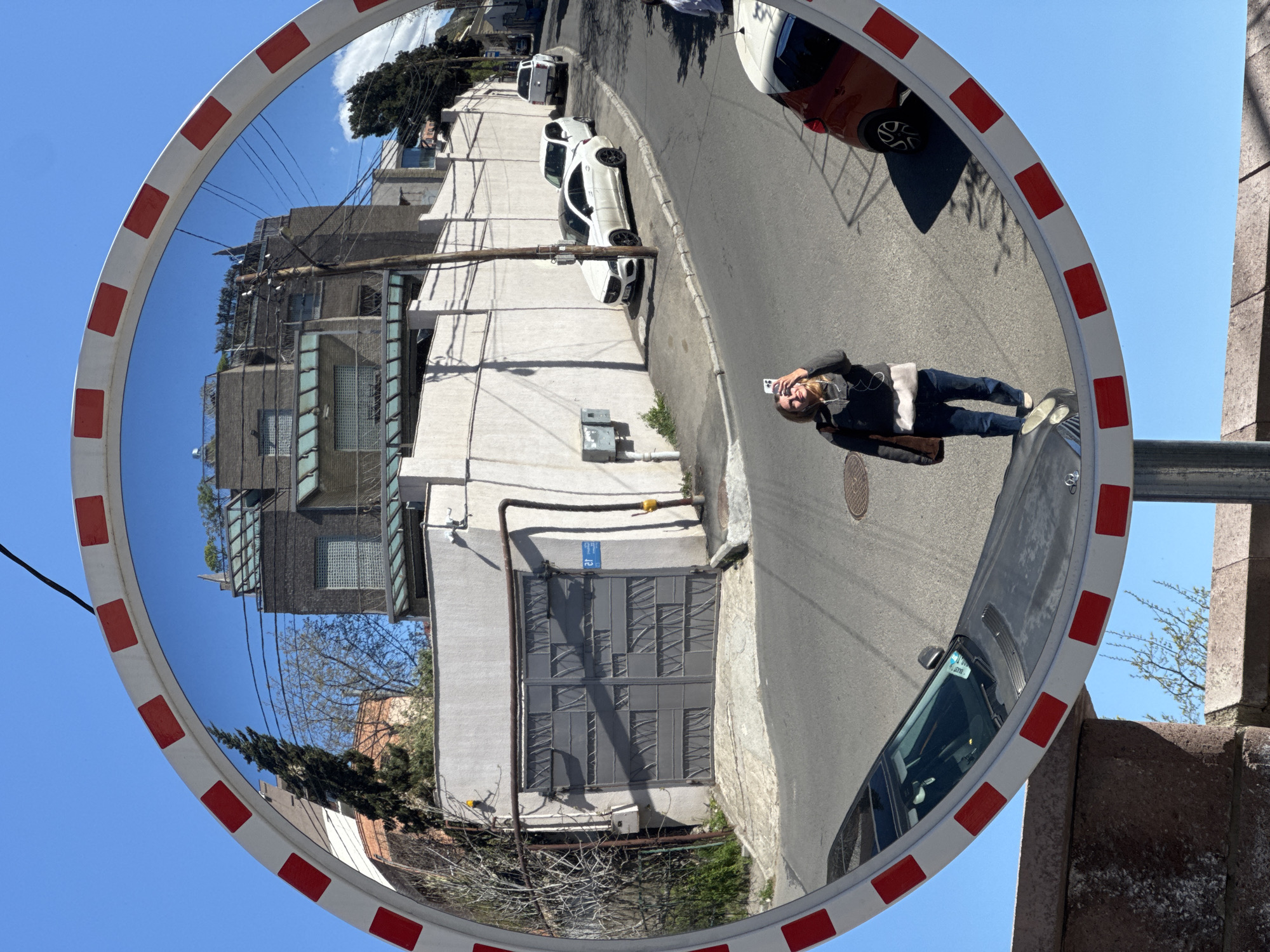 The image is a reflection in a convex security mirror on a street in Tbilisi, Georgia. The mirror reflects a street scene with several parked cars, including a white sedan and an orange vehicle. A building with a gray gate and utility lines is visible. A person is also reflected in the mirror, holding a phone and seemingly taking a picture. The mirror has a white frame with alternating red rectangular markers. The sky is blue.