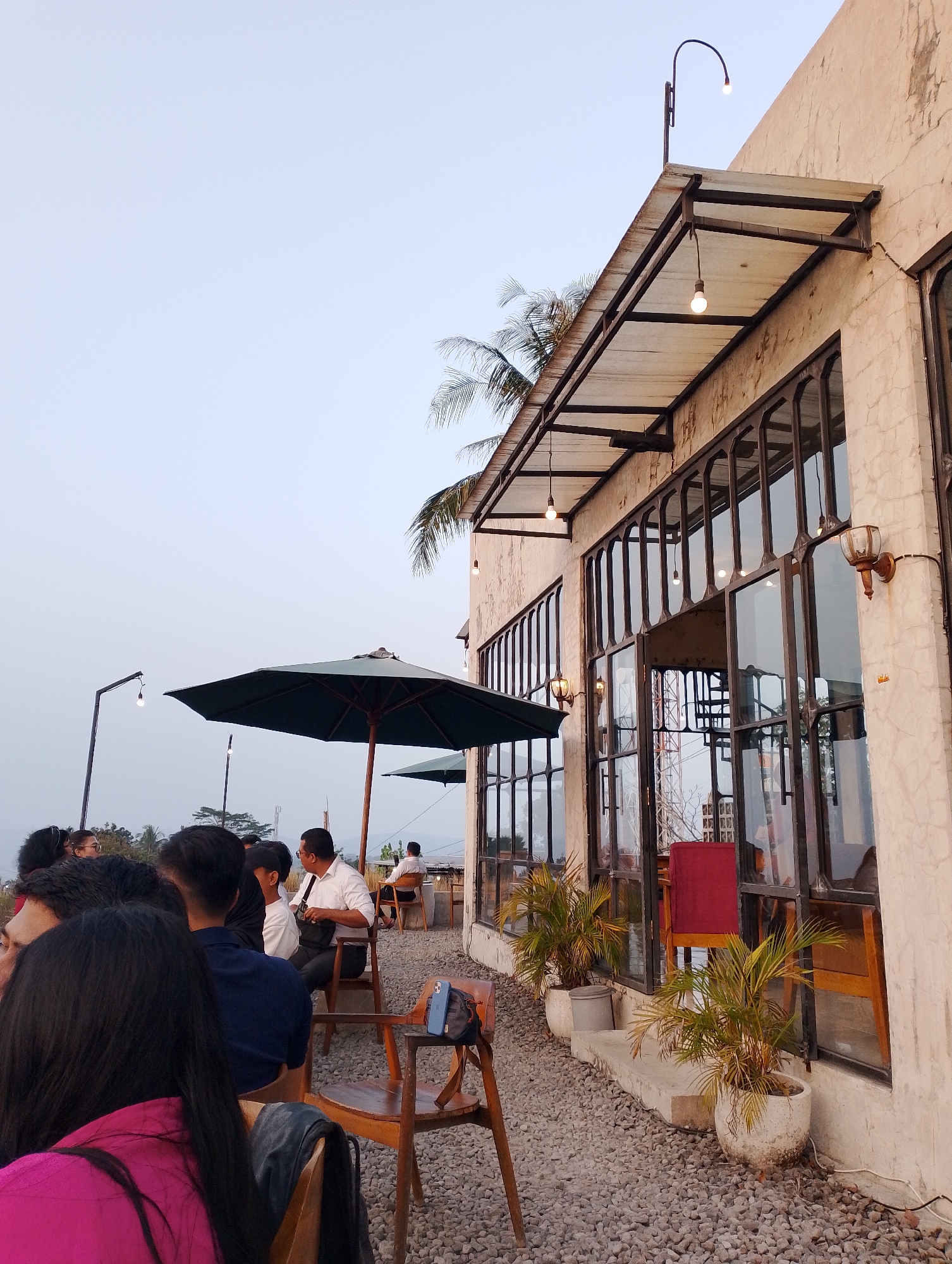 The image depicts an outdoor seating area of a cafe in Srimulyo, Indonesia, as suggested by the context. The cafe has a rough, textured light-colored exterior wall and large, dark-framed windows. An awning supported by metal beams extends over a section of the seating area. Multiple light bulbs are visible, some hanging from wires and others attached to the wall as sconces. The ground is covered in gravel.
Several people are seated at tables, some under a large dark green umbrella. The individuals appear to be customers, engaged in conversation or observing their surroundings. One man in a white shirt is sitting at a table, facing away from the camera. In the foreground, a woman with long black hair wearing a bright pink top is partially visible. Other patrons are scattered throughout the seating area. Two potted plants with spiky fronds are placed near the windows. The sky is a pale, overcast blue. In the background, distant hills and trees are visible, suggesting a semi-rural or elevated location.
